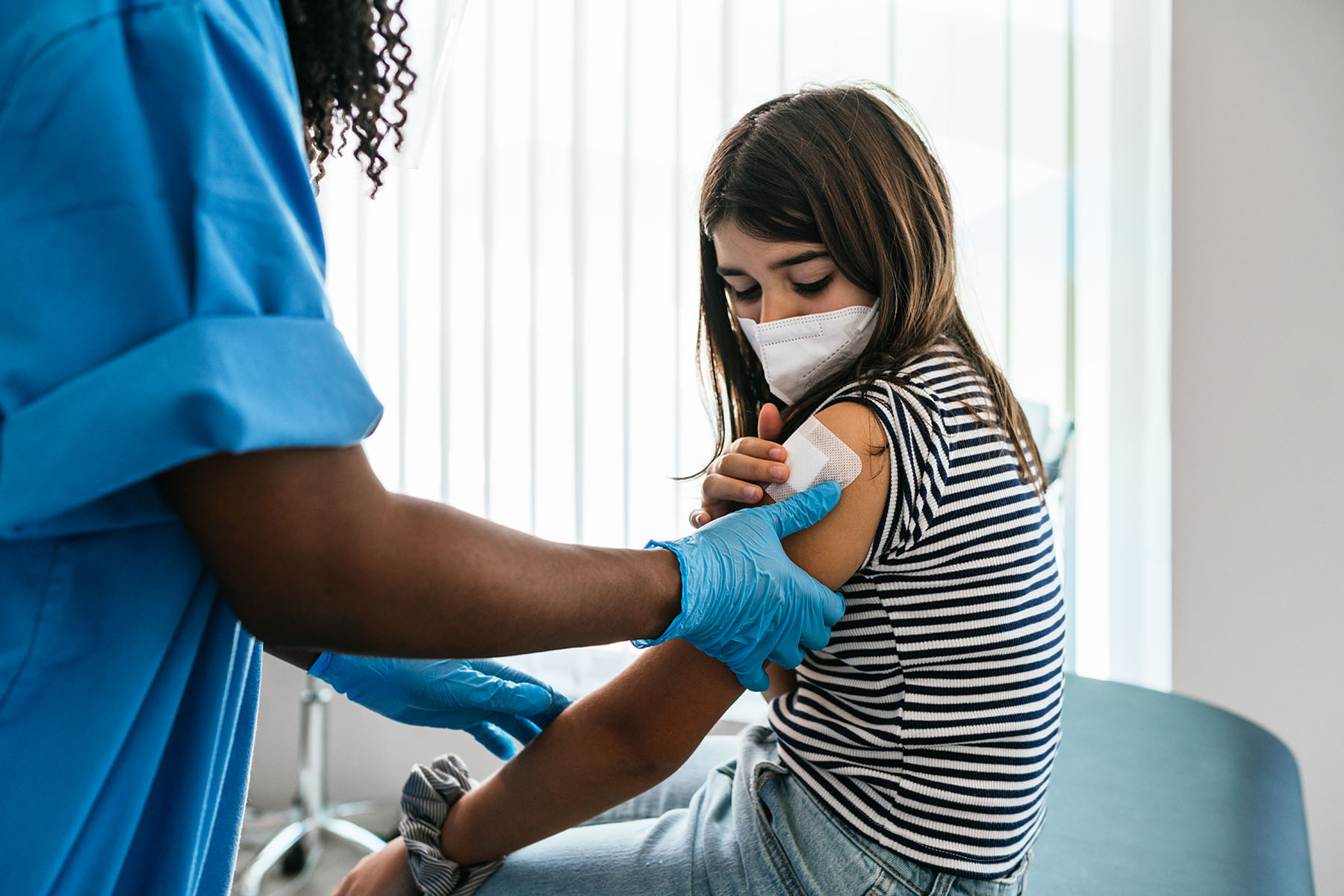 Medical health professional putting a bandage on a girls arm, after she received a vaccination. The girl is wearing a face mask.
