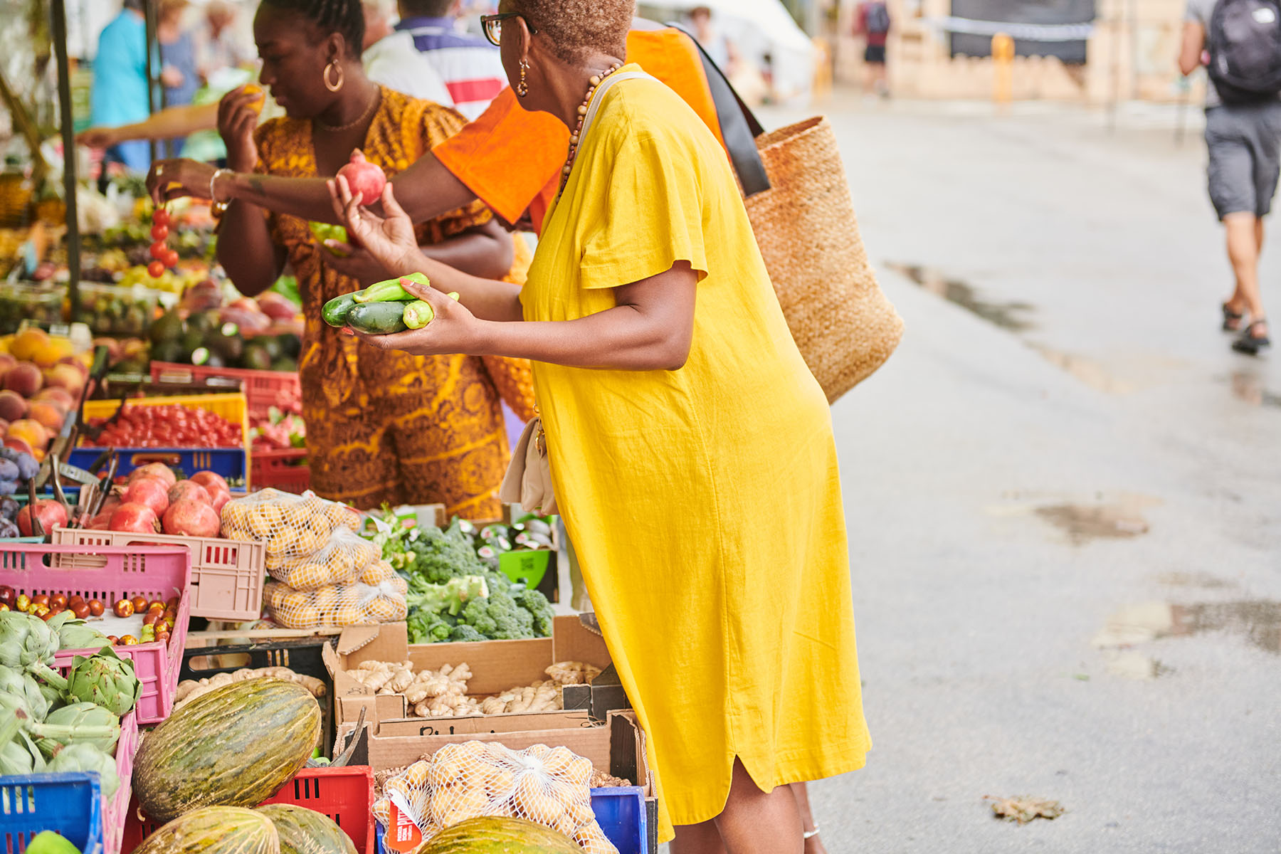 Woman in a yellow dress looking at a pomegranate and cucumbers at a veggie market stall.