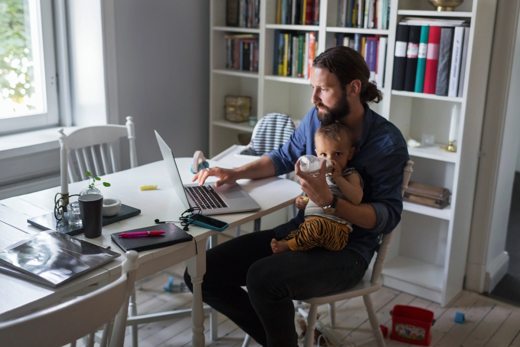 a father feeding his baby a bottle on his lap as he works on his laptop in his home office