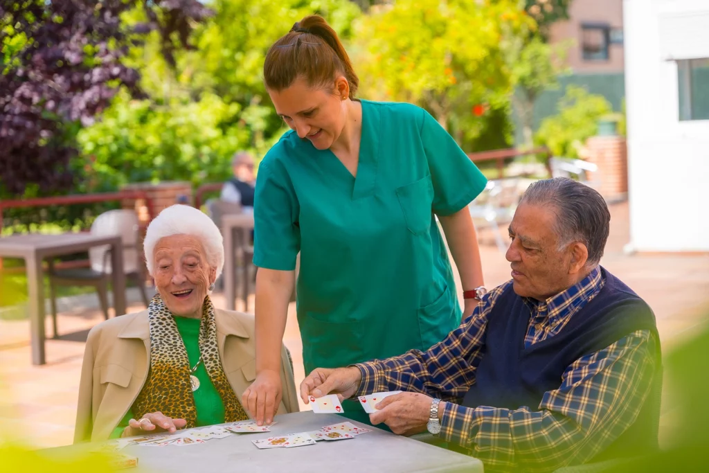 A senior couple plays cards while their retirement home's nurse looks at the cards and smiles - services can be covered by health insurnace for seniors in Spain