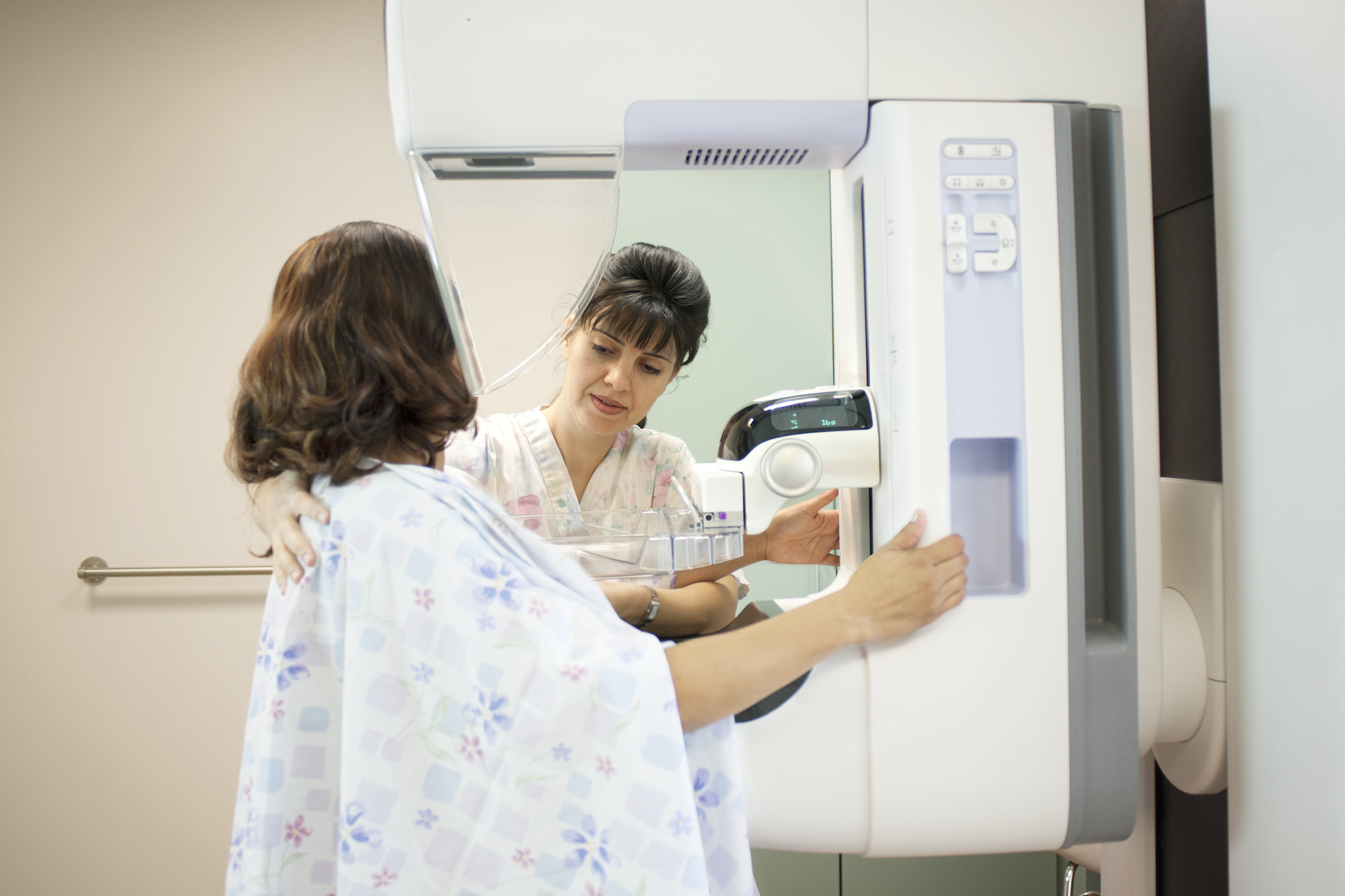 A nurse performs a mammogram, the patient is holding onto the imagery machine with head turned away from the camera