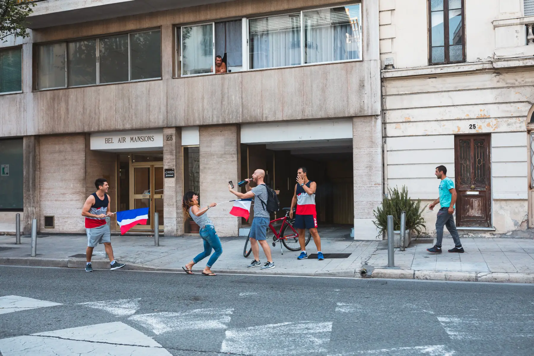 People waving French flags and celebrating on the sidewalk outside an apartment building. 