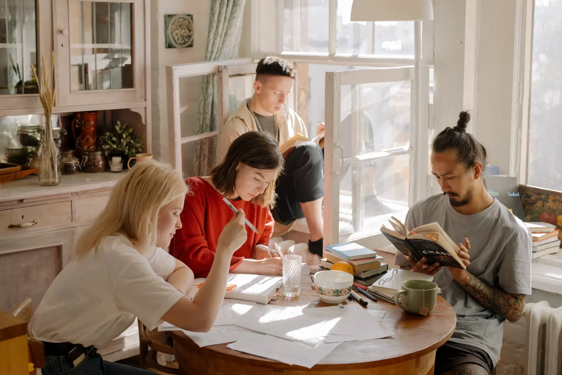 A group of students, (two women and one man) studying at a round table. One man is sitting in the window sill reading a book.