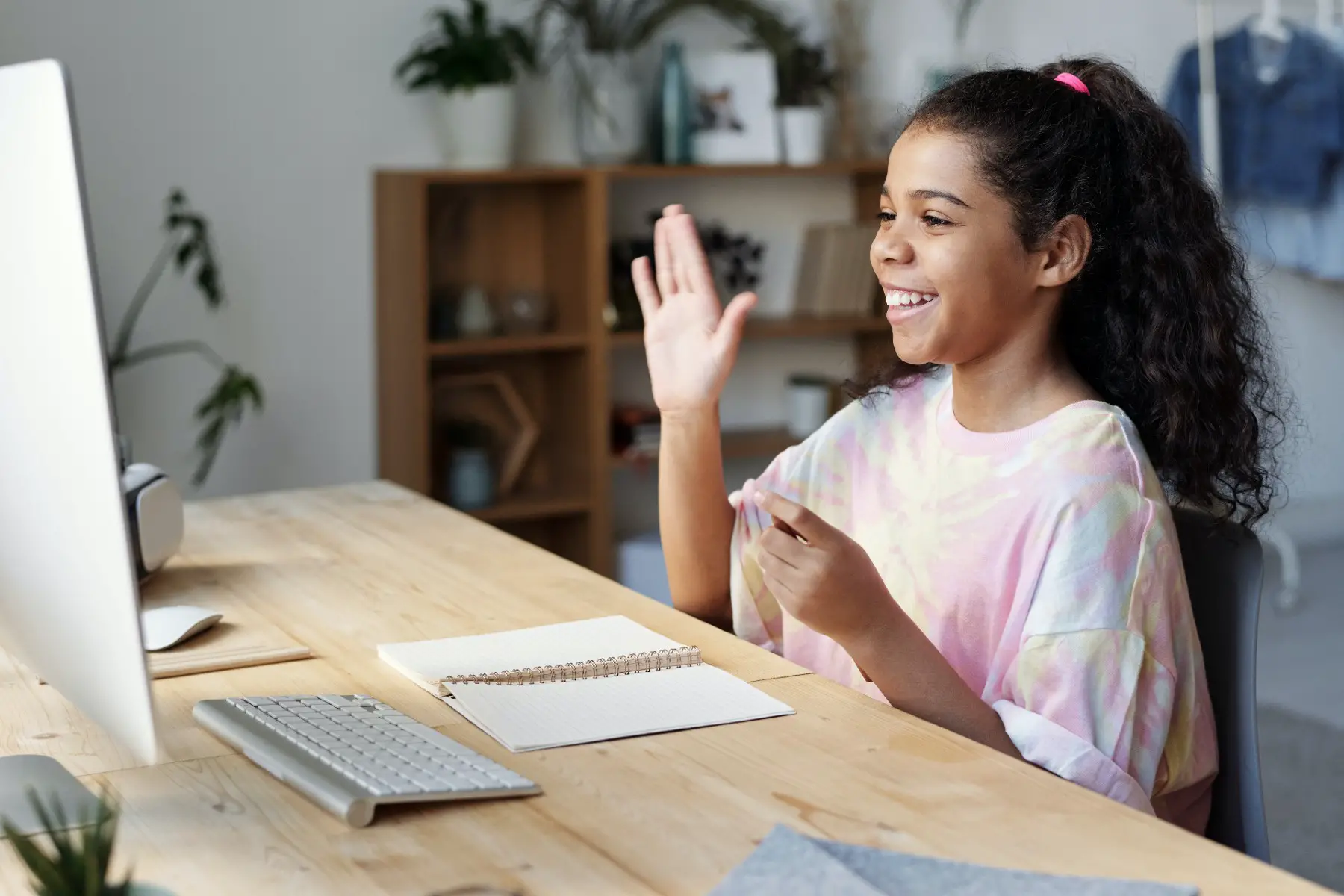 School-aged girl smiling and waving hello to a computer screen.