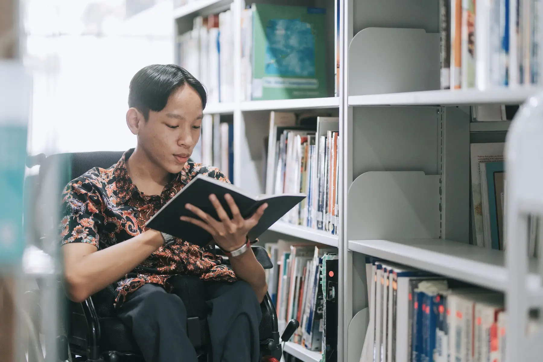 Student living with a disability in a wheelchair reading book in library.
