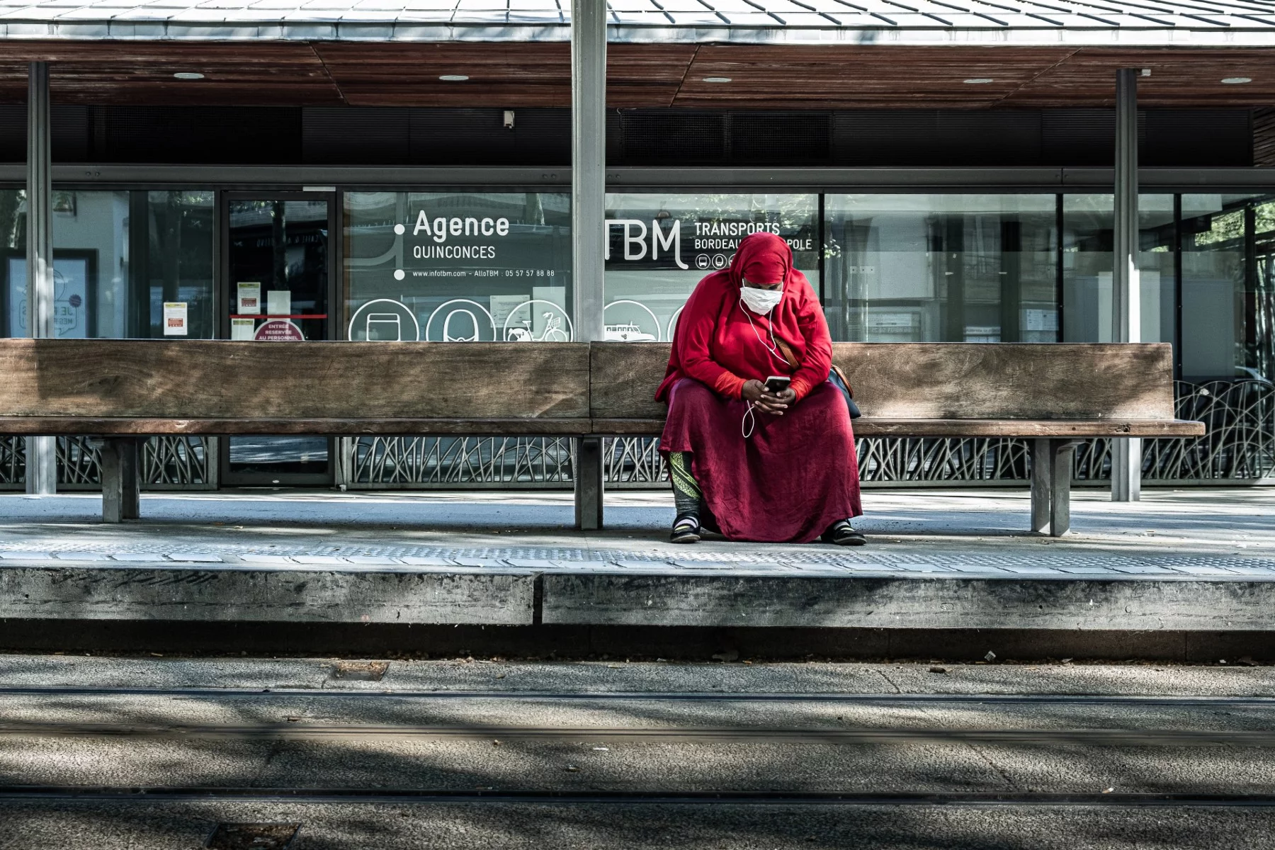 A lady in a red burqa, listening to music on her phone as she waits on public transport. 