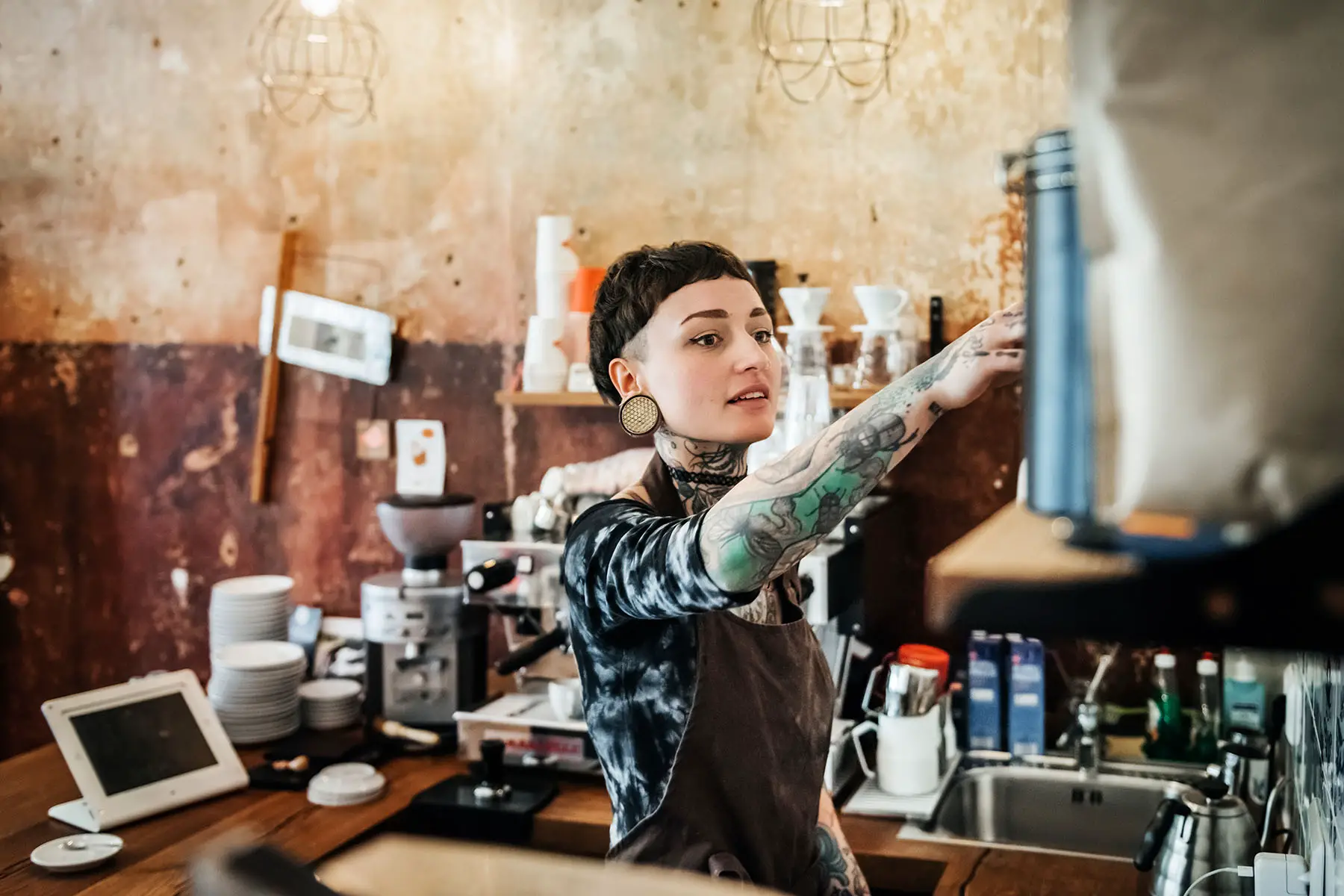 A young tattooed woman wearing an apron is working as a waitress in a bright café.