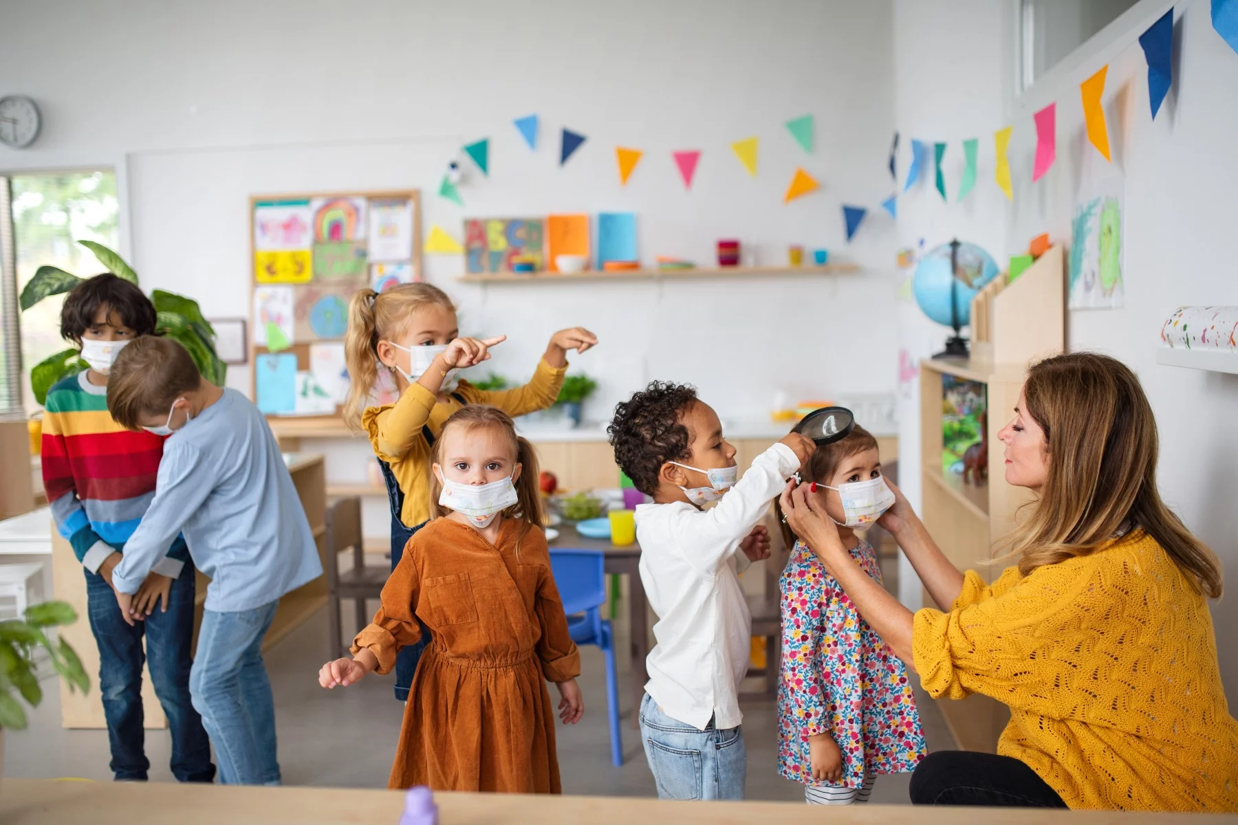 Preschool teacher helping little girl put on a face mask indoors at nursery school.