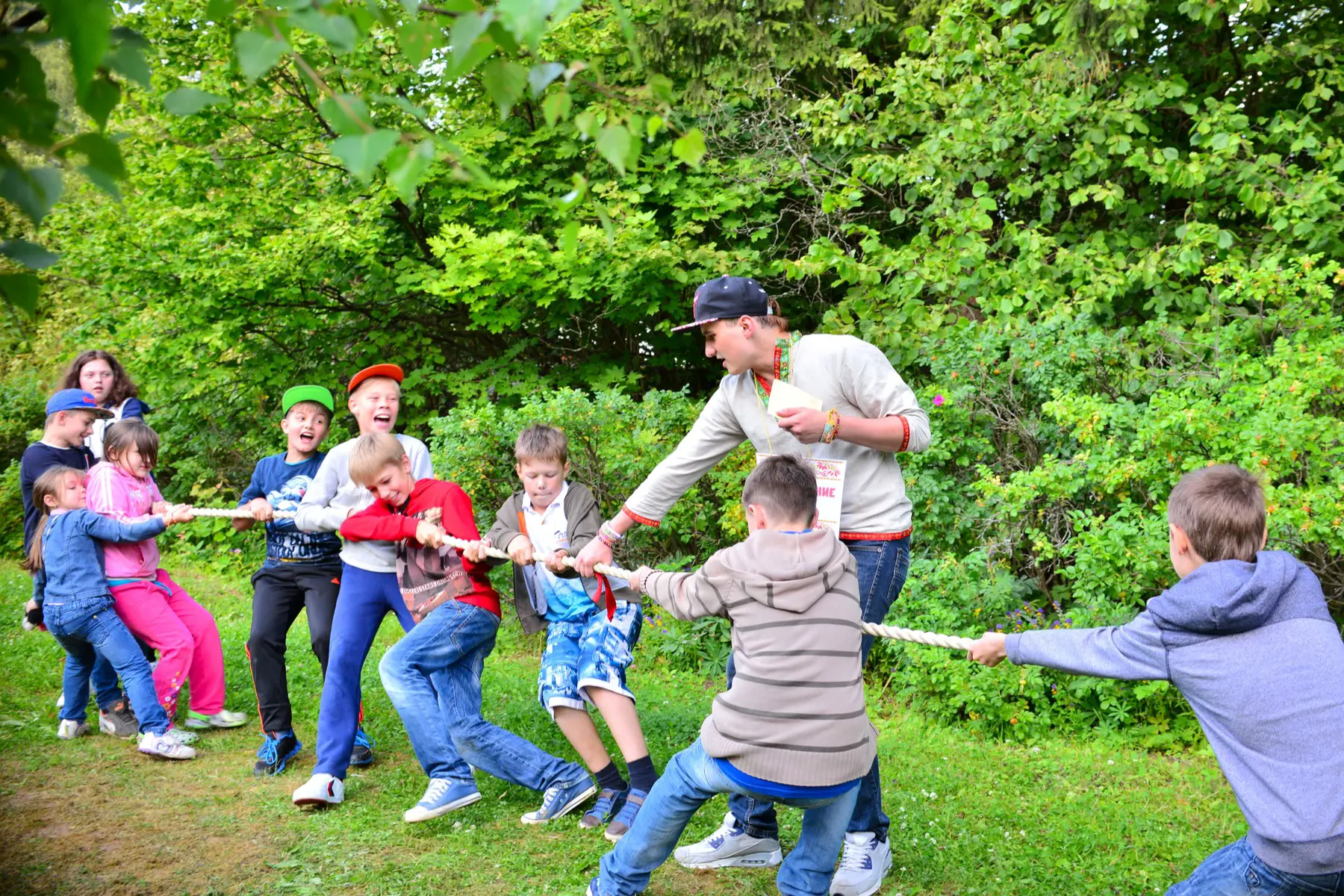 children play tug of war amid greenery as a young man supervises them
