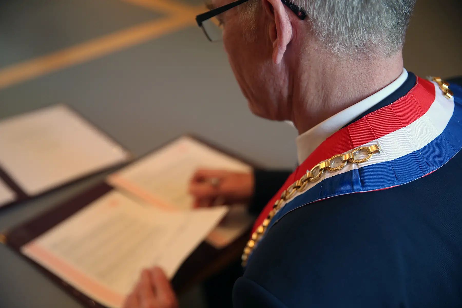 View of a mayor from behind, wearing a French-flag sash and chain, looking at documents on a table at a civil wedding ceremony