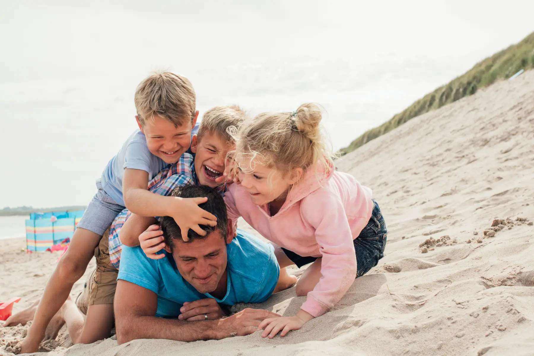 three children having fun at the beach with their father, pinning him down and tickling him