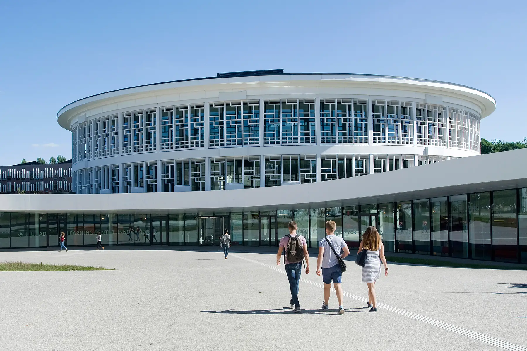 Students walking outside a large, white, cylindrical, modern university building on a sunny day.
