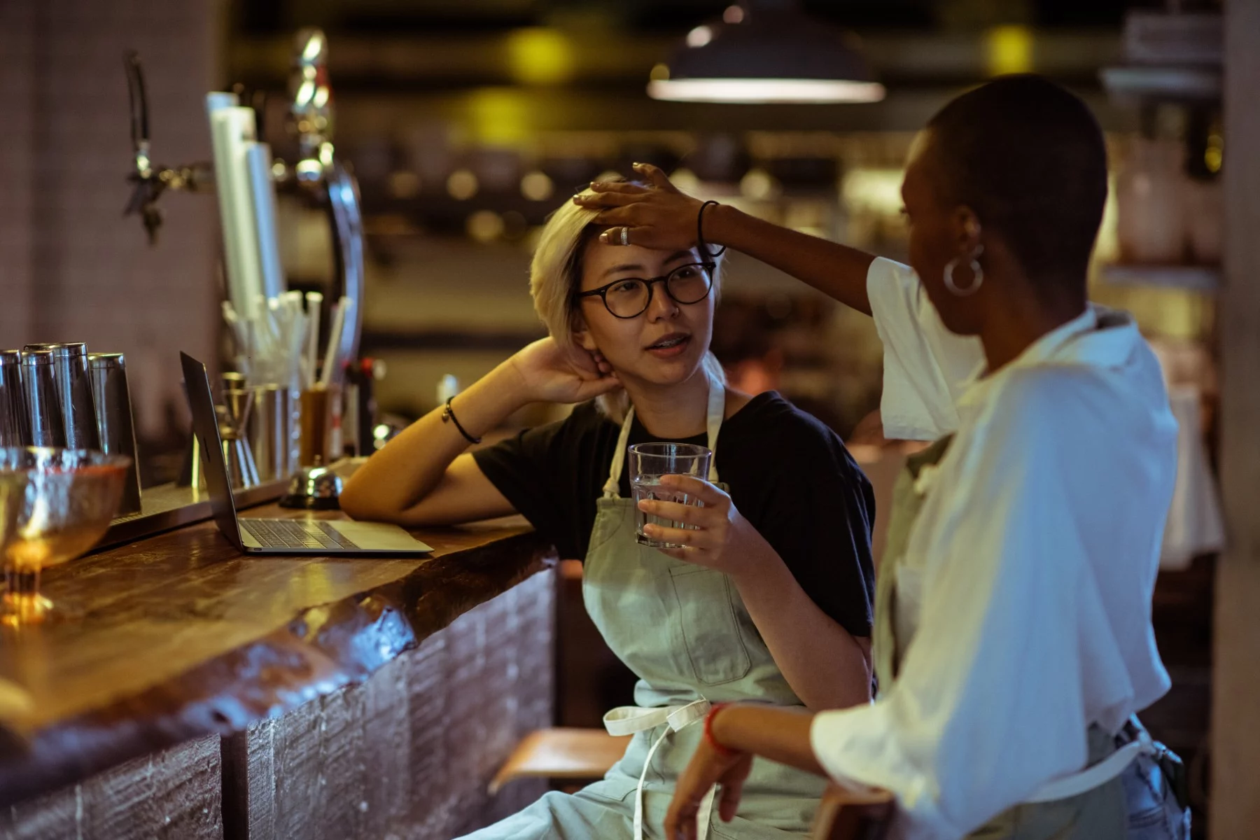 Female worker feeling the forehead of another female worker in a restaurant, sitting at the bar.