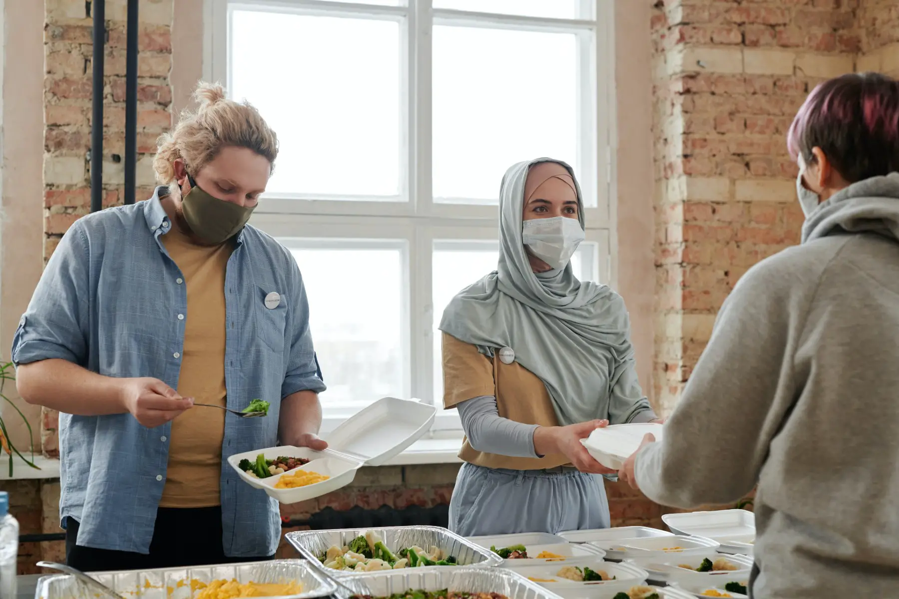 A person plating food in a plastic container, while a woman with a headscarf hands out food.