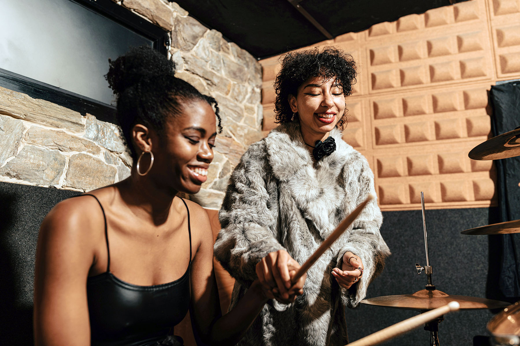 Two female musiscians laughing as they're playing drums in a recording studio. One is wearing a massive fur coat.