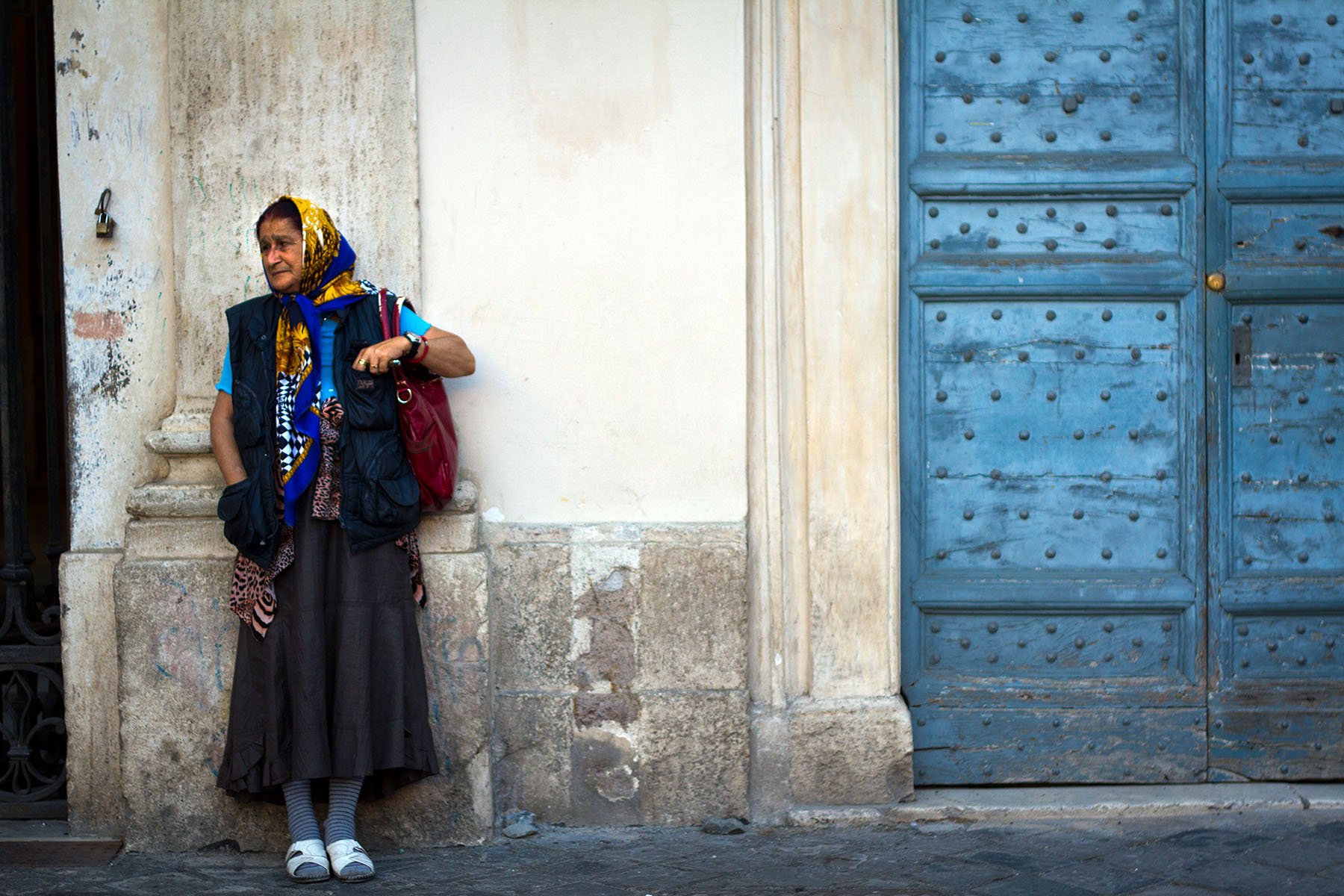 A Roma woman is standing on the street, next to large blue doors of the Basilica of Santa Maria in Rome's Trastevere district. She's colorfully dressed, but looking sad.