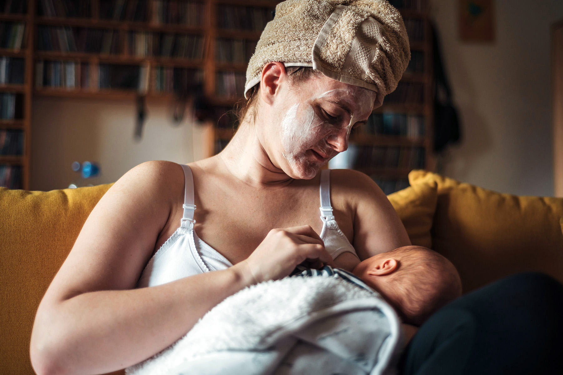 Woman sitting on a yellow couch at home, she's wearing a moisturizing face mask and has her hair wrapped in a towel. She's looking down at the newborn baby in her arms, who is breastfeeding.