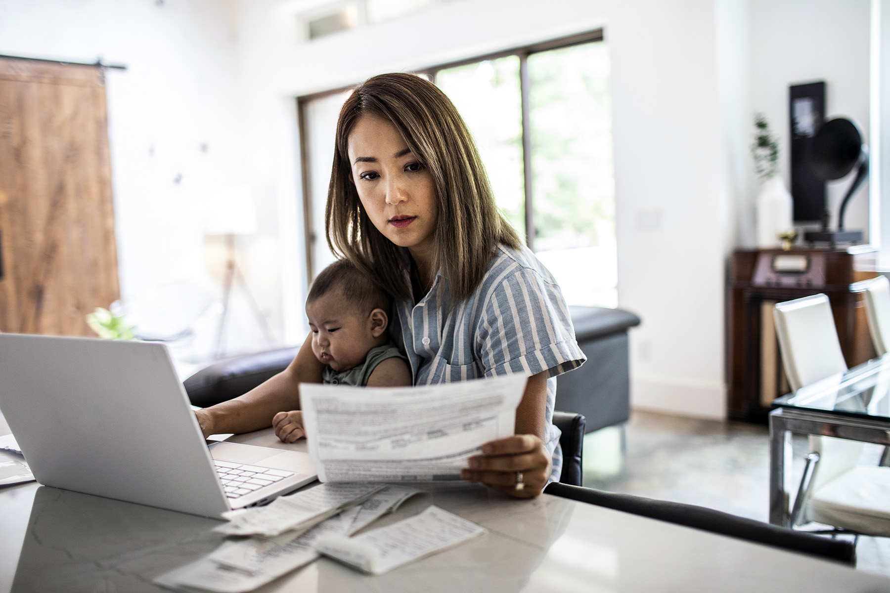 Mother working from home while holding baby on her lap. She's looking at a document she's holding in her hand.