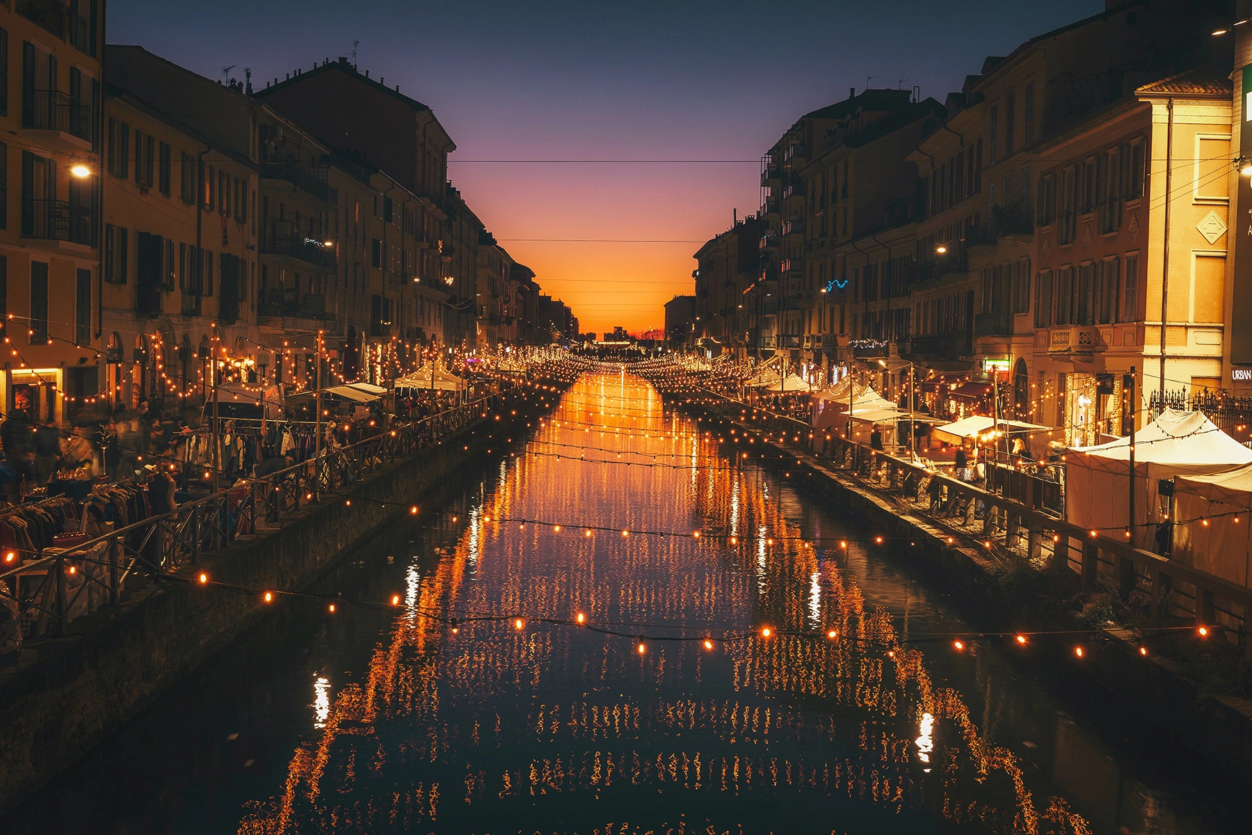 Alzaia Naviglio Grande in Milan, Italy. Lights hang over a canal in the evening while people dine outside.