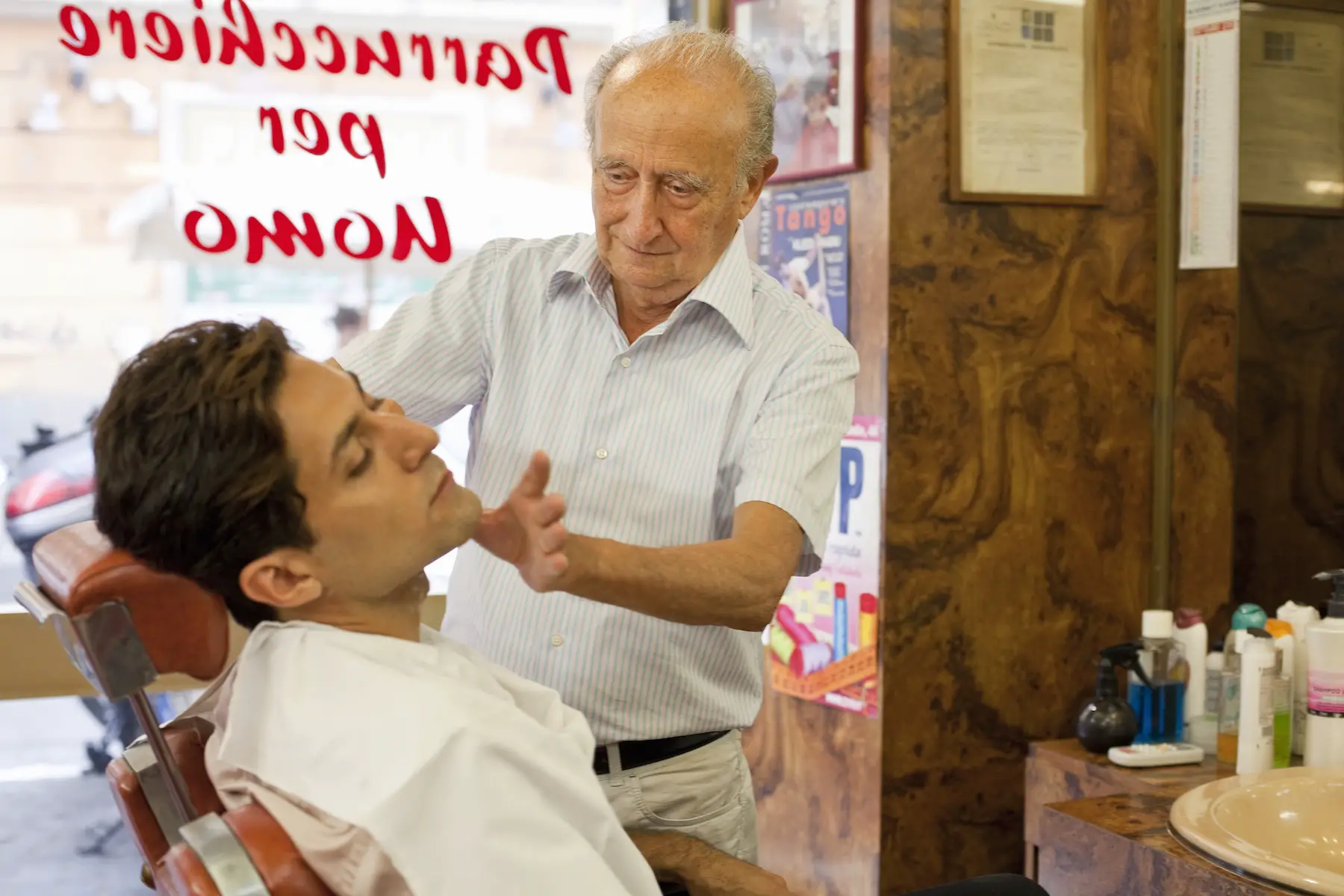 A young man gets a traditional shave from an older man at a classic barber shop in Rome; his professional license is displayed on the wall.