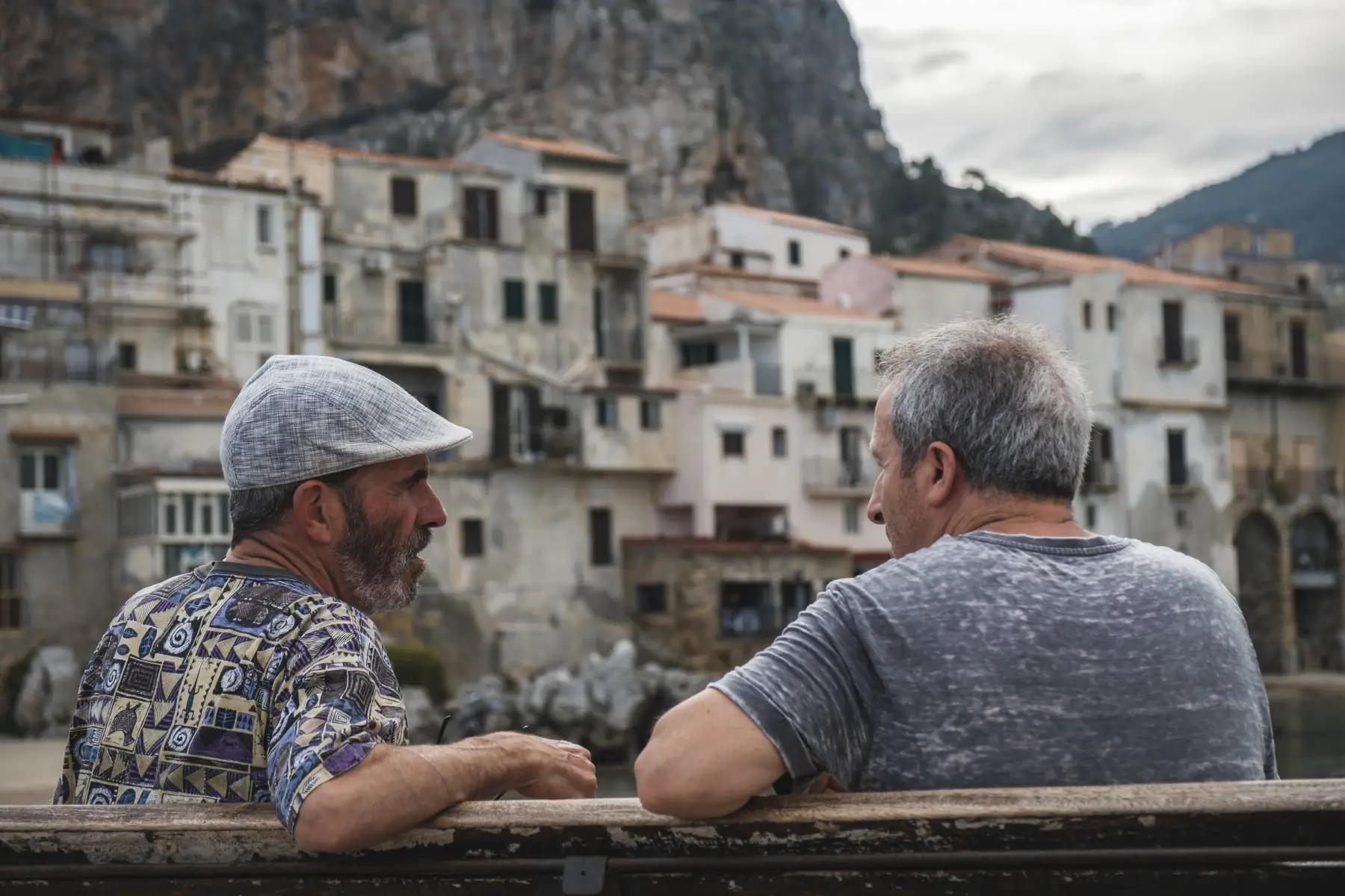 Two men having a deep conversation on a bench, the village of Cefalu in the background