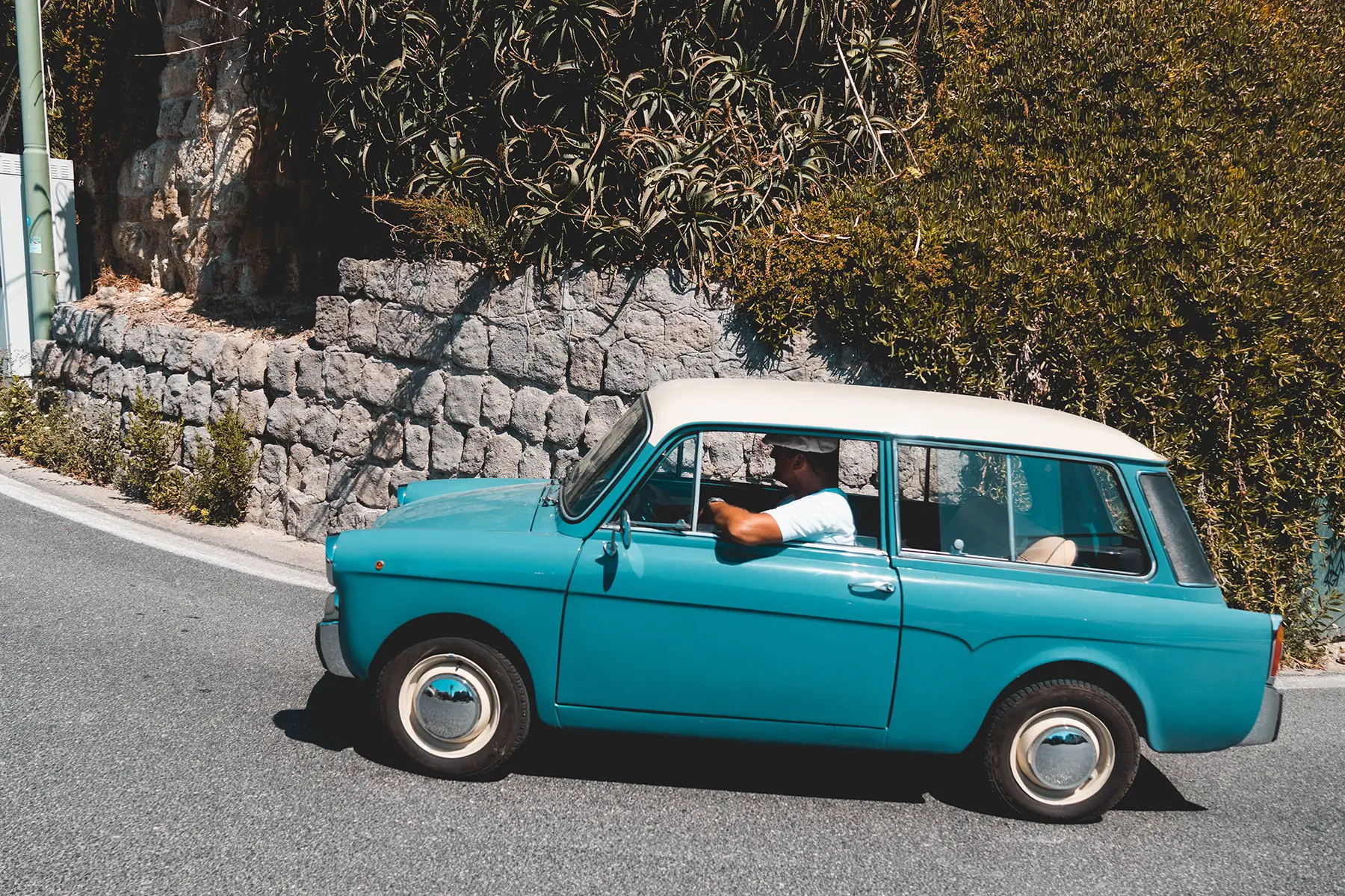 A man driving a blue Autobianchi Bianchina with a white roof up a hill.