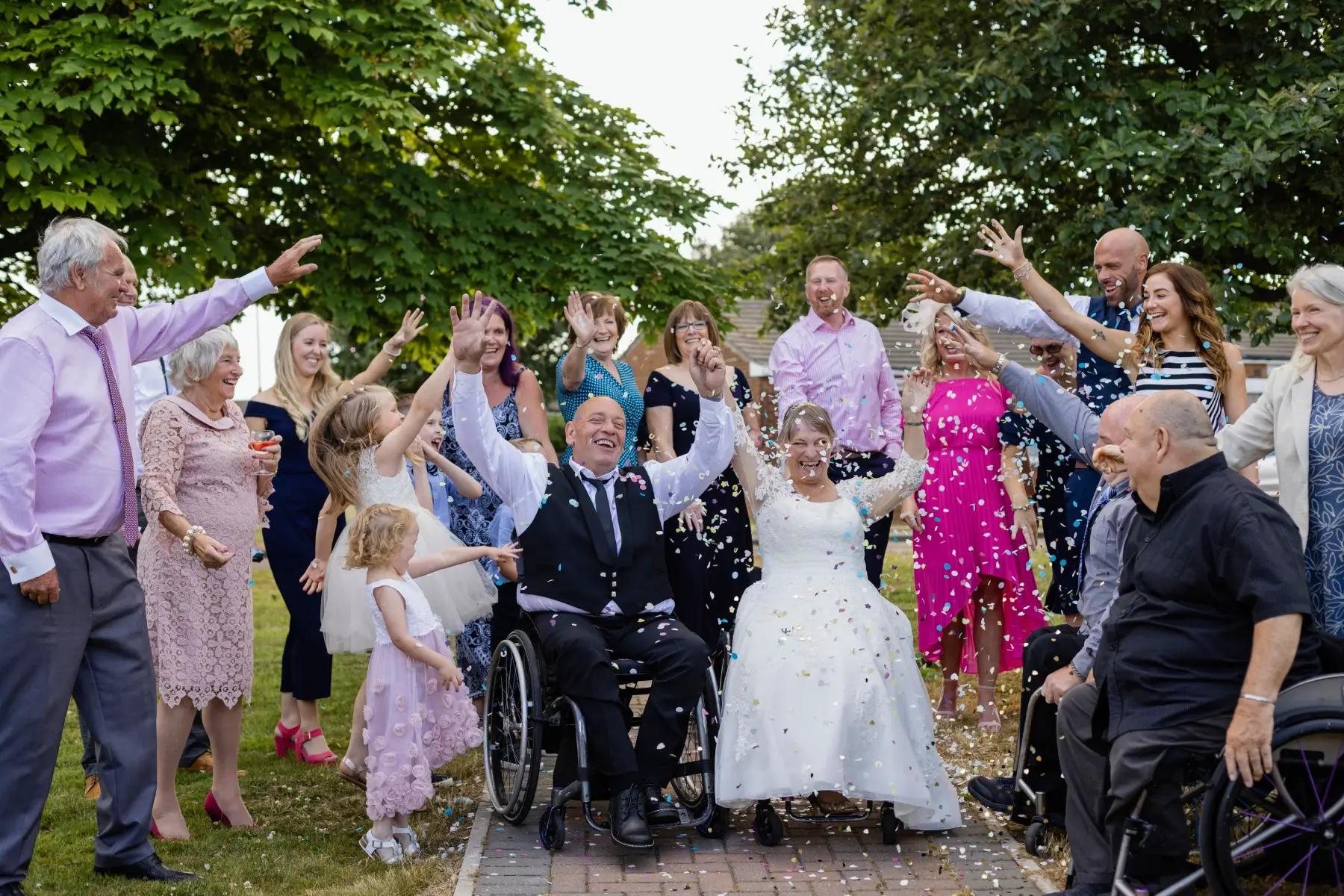 Friends and family throw confetti on a bridal couple seated in their wheelchairs