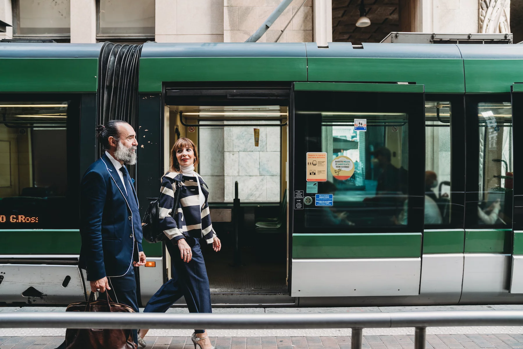 A smartly dressed man and woman walk alongside a green tram in Milan on their way to work