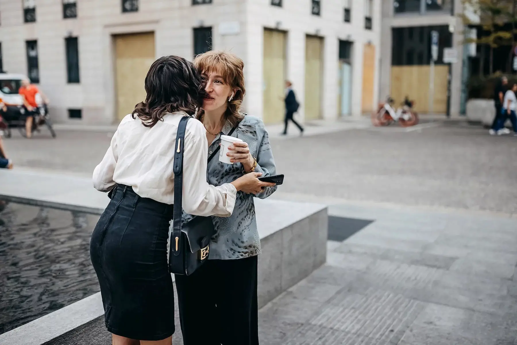 Two well-dressed businesswomen meet on the city street, first kissing each other's cheek