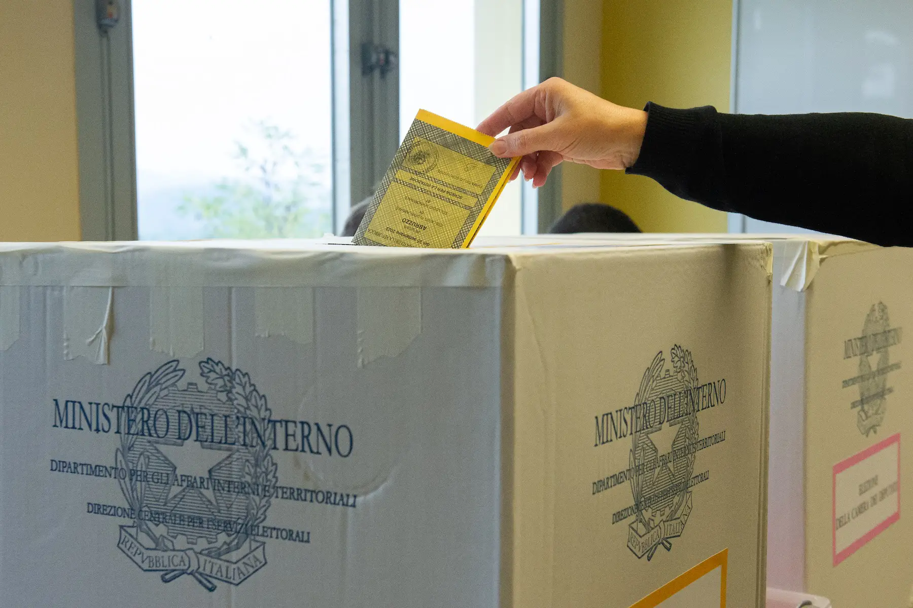 A hand drops a bright yellow voter ballot into the cardboard ballot box during an Italian election