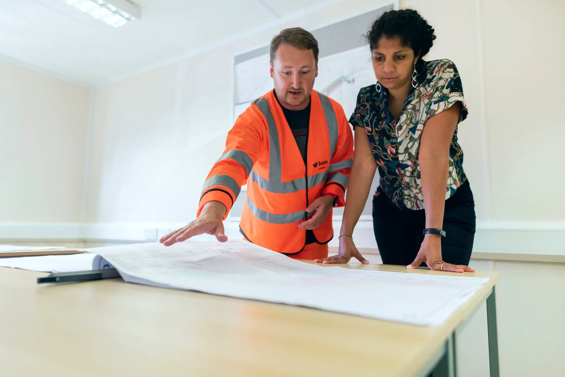 A man and woman standing over a table looking at a civil engineering work plan. He wears a construction jacket and she wears business attire.