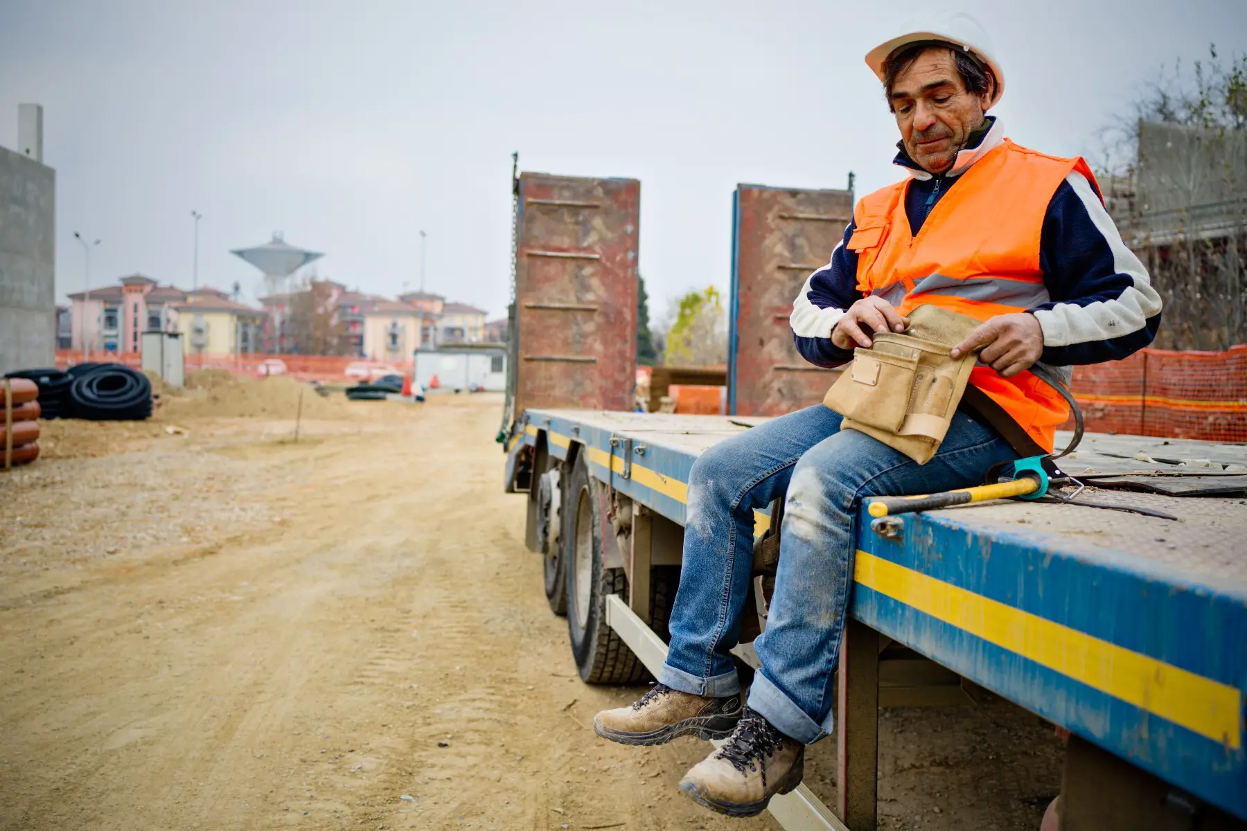 A construction worker in an orange vest and hard hat takes a break sitting on the bed of a trailer truck