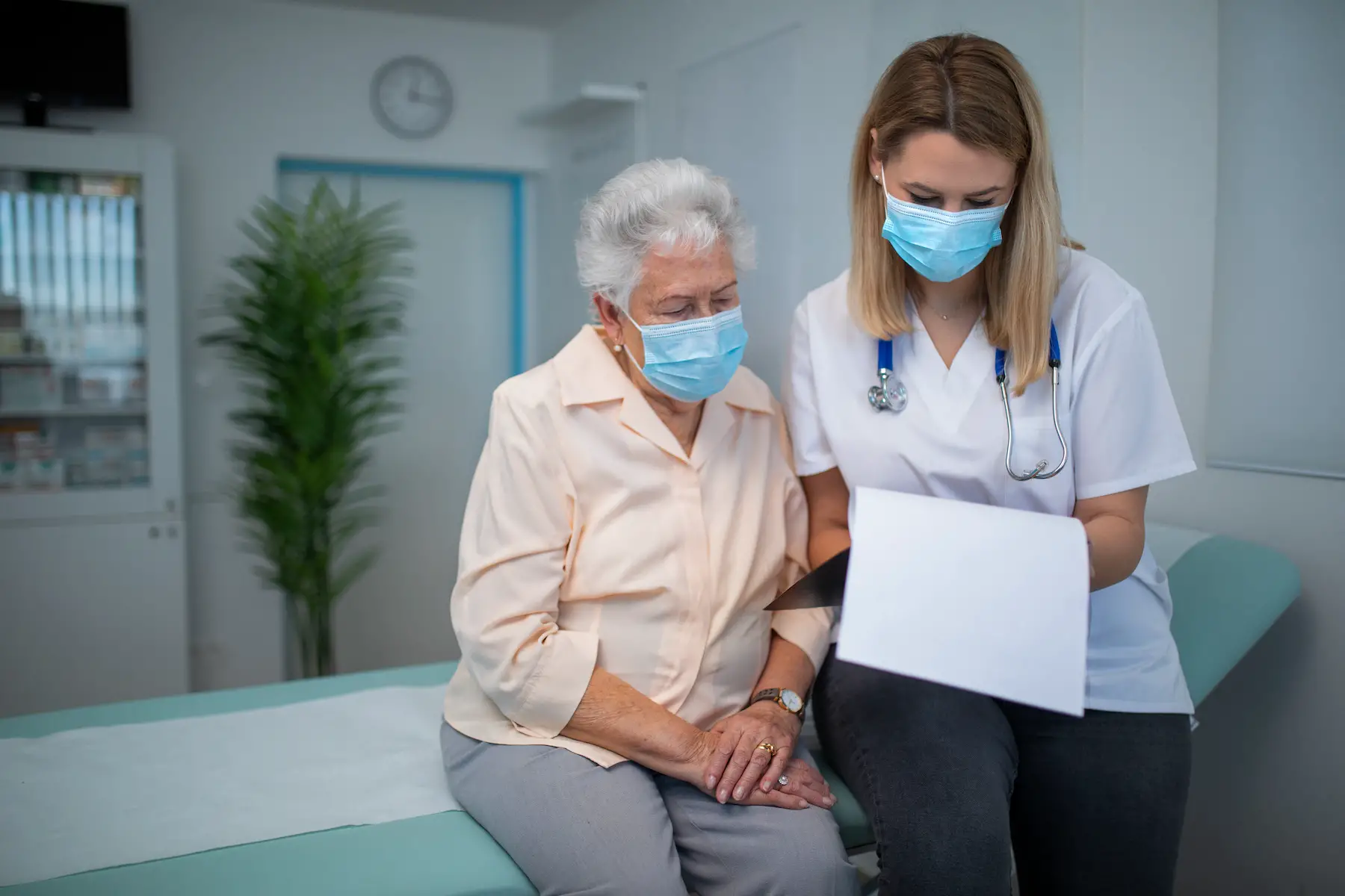 A doctor sits with her senior patient, looking over her chart together; they are both wearing masks