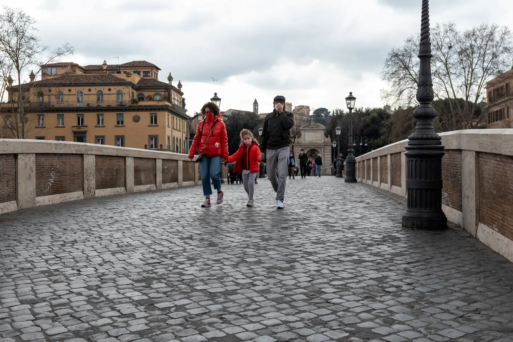A family walking across a cobbled bridge on a cloudy day, wearing face masks.