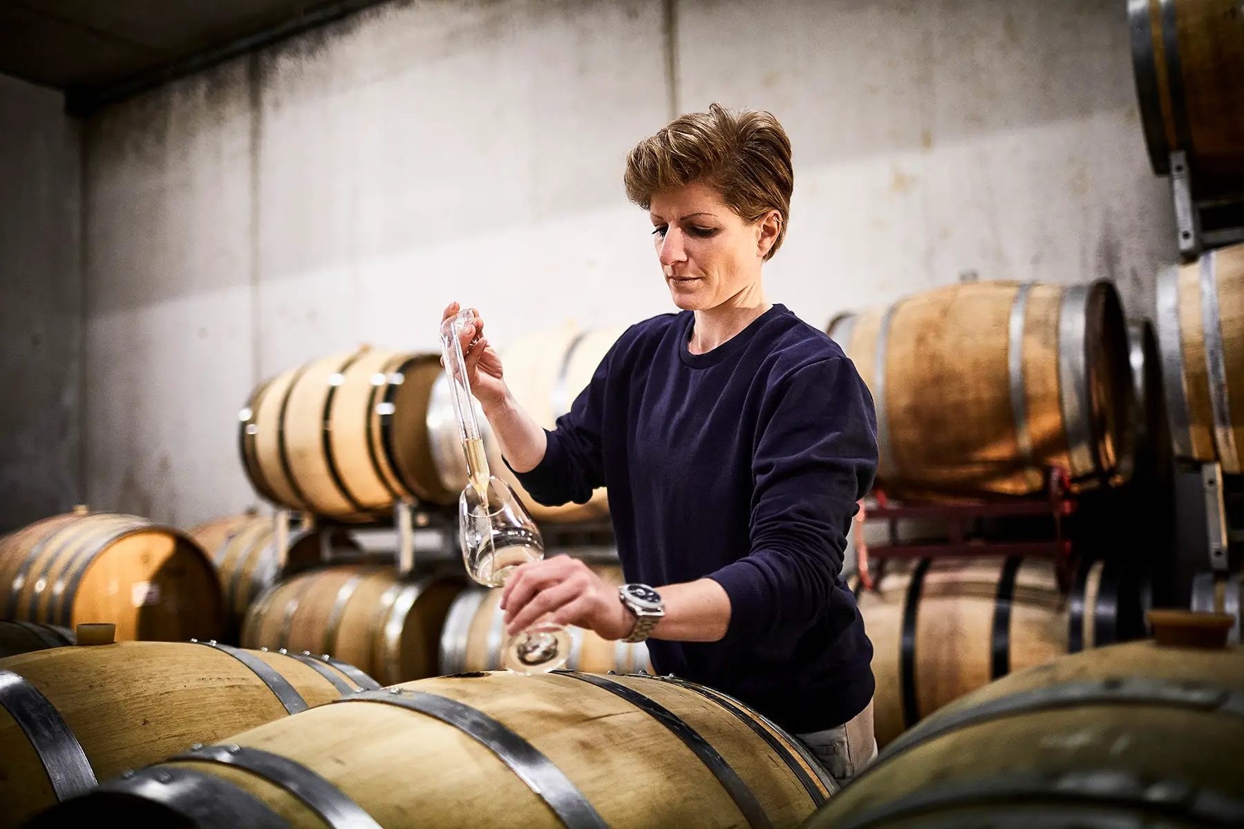 A vintner (woman) testing the wine she made, standing between wooden barrels in a cellar