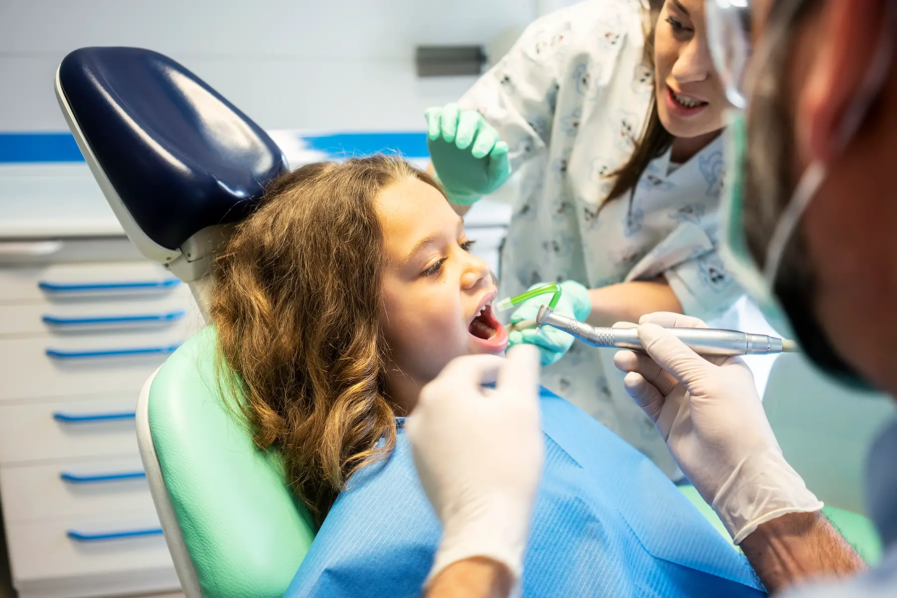 A girl in a dentist's chair having various dental instruments placed into her mouth by two dentists