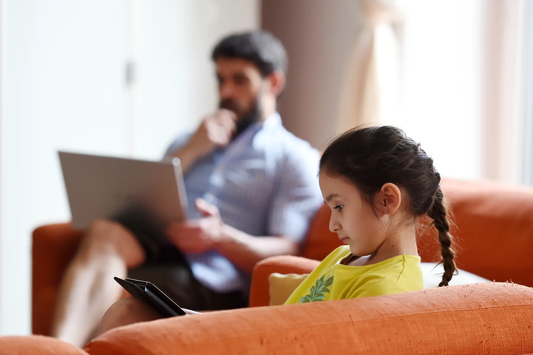 A girl in the foreground sits on the sofa and is looking at a tablet. In the background, out of focus, her father works on a laptop.