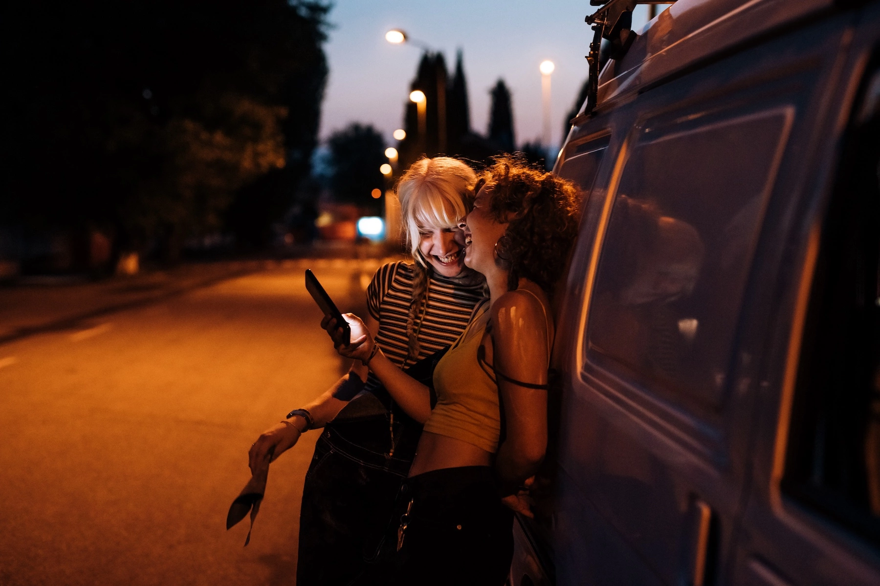 Two young female friends lean against a van and laugh watching a video on a mobile phone