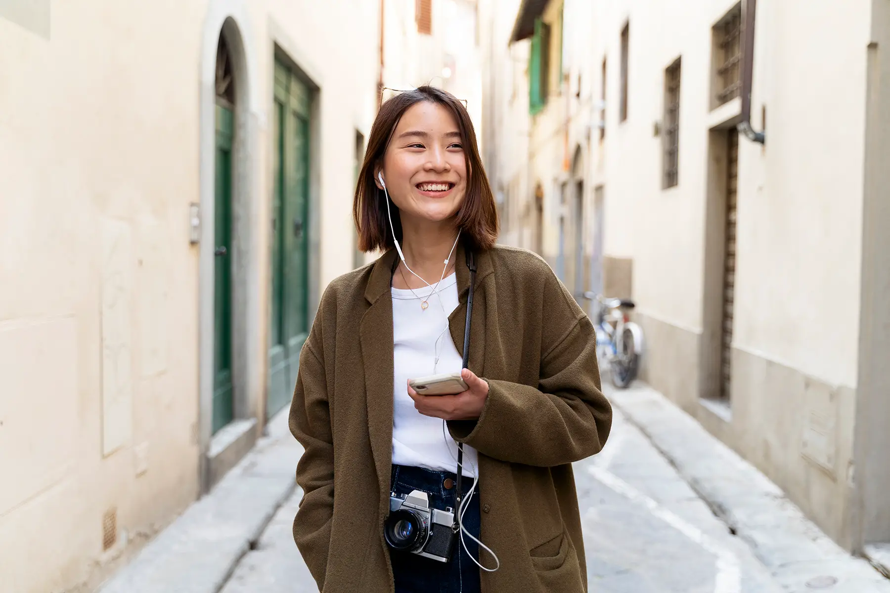 A happy woman walking down an Italian street holding a phone. She is wearing headphones that are connected to the phone.