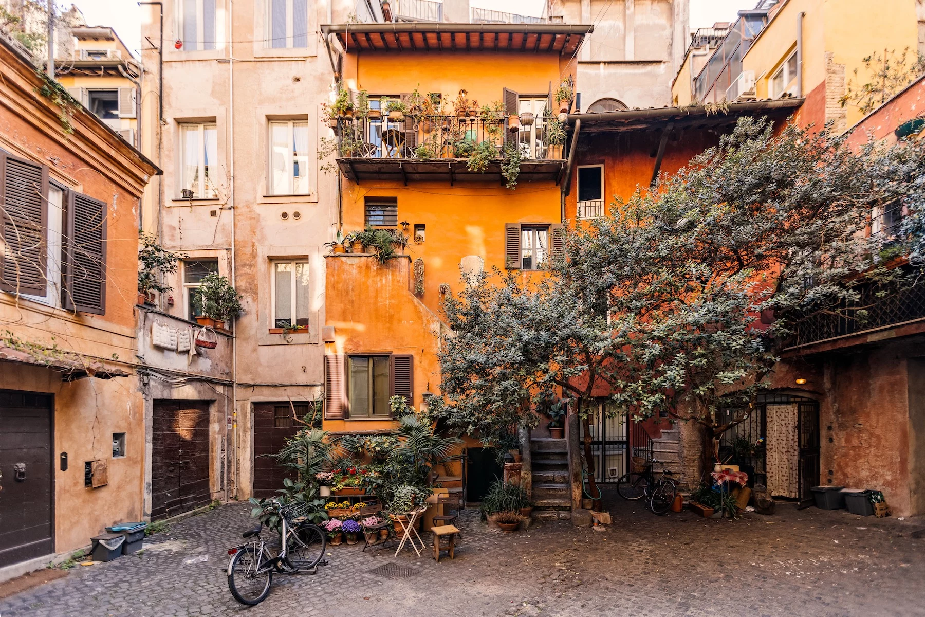 Old Italian apartment buildings create a sunny courtyard full of trees, flowers, and bicycles