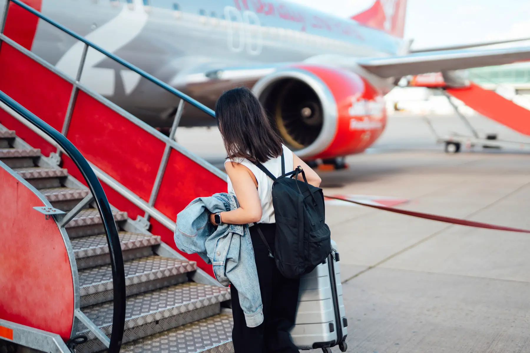 Young woman with suitcase, walking on the runway towards the stairs to the airplane.