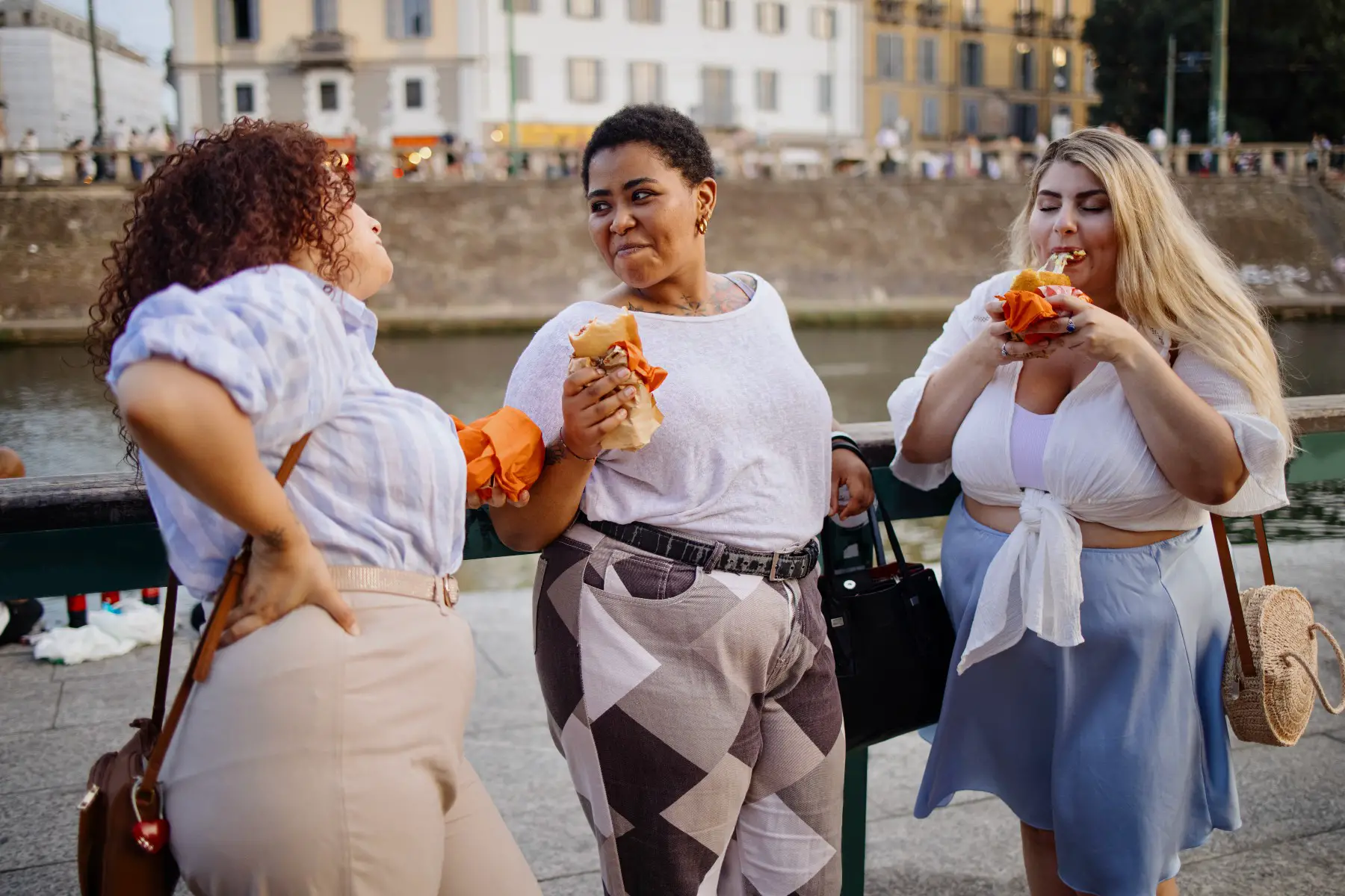 Three friends enjoying a snack on the go in the Navigli District in Milan, Italy. The blond woman is thoroughly enjoying an arancini.