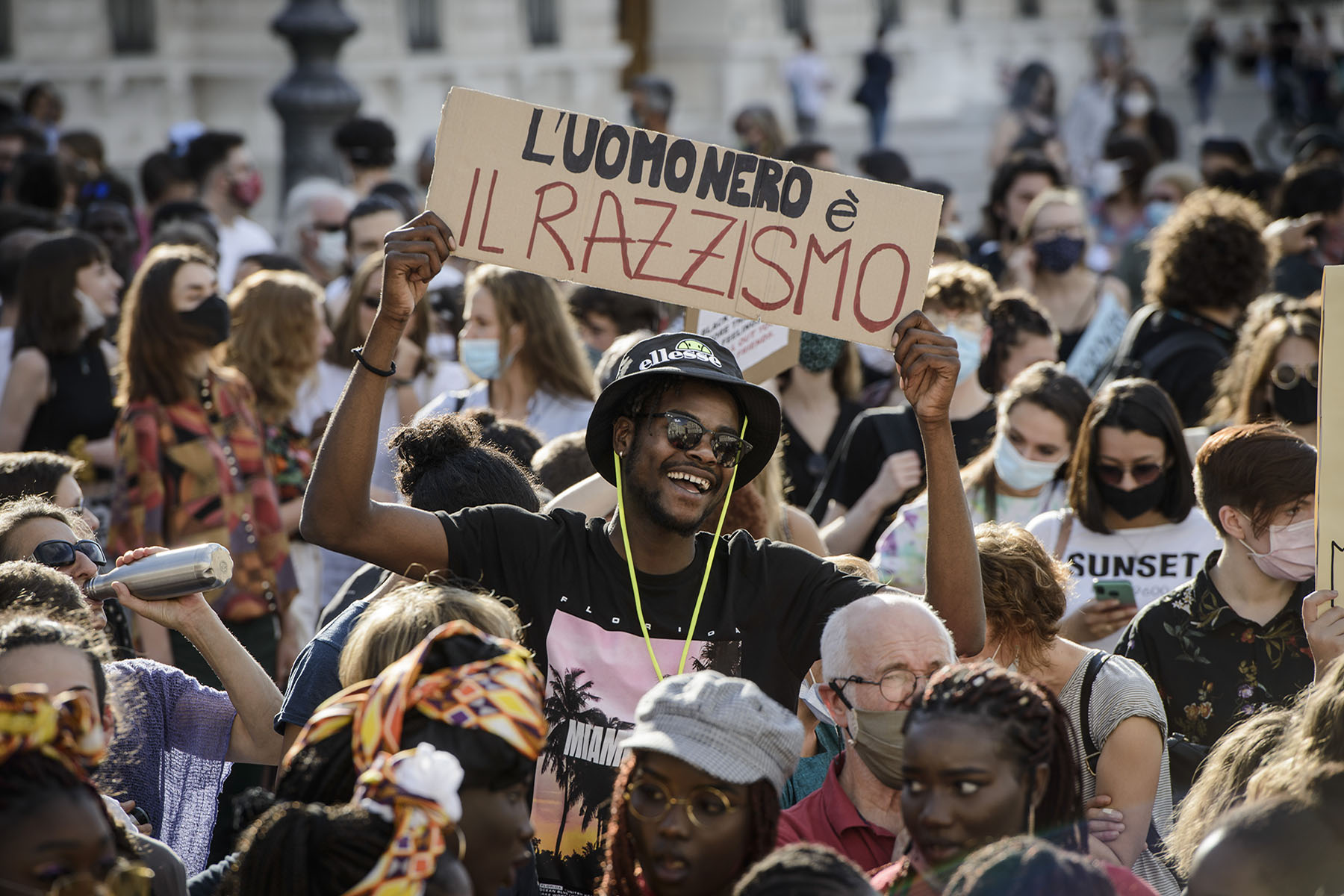 Protesters gathering with signs and messages to celebrate Black Culture during the 2020 BLM Protest in Trieste, Italy. Photo is focussed on a smiling Black man holding a sign that says 