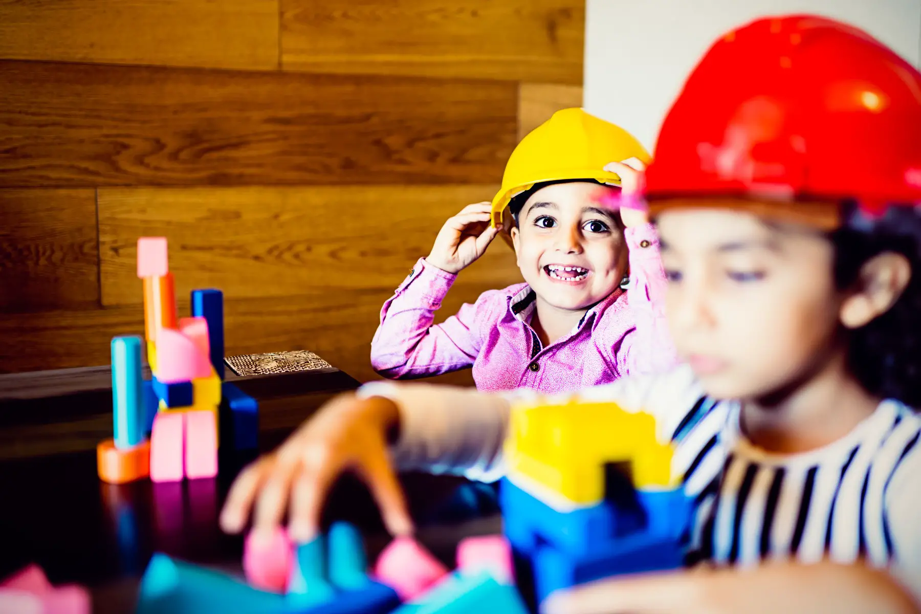 Children playing with building blocks, and wearing builders helmets. One cute kid is looking directly into the camera and smiling.