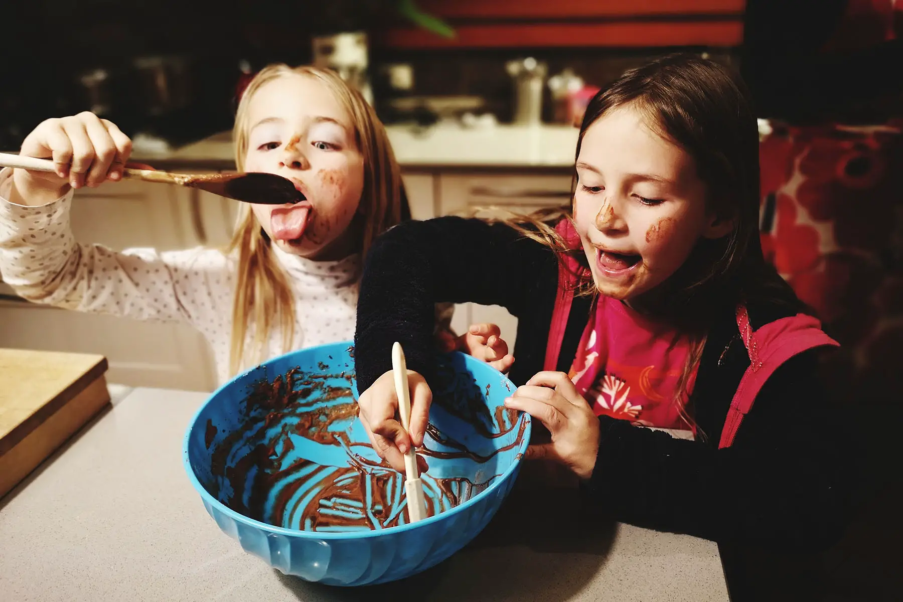 Two sisters using a spoon to get the last bit of chocolate cake batter out of the bowl. They are covered in chocolate mix.