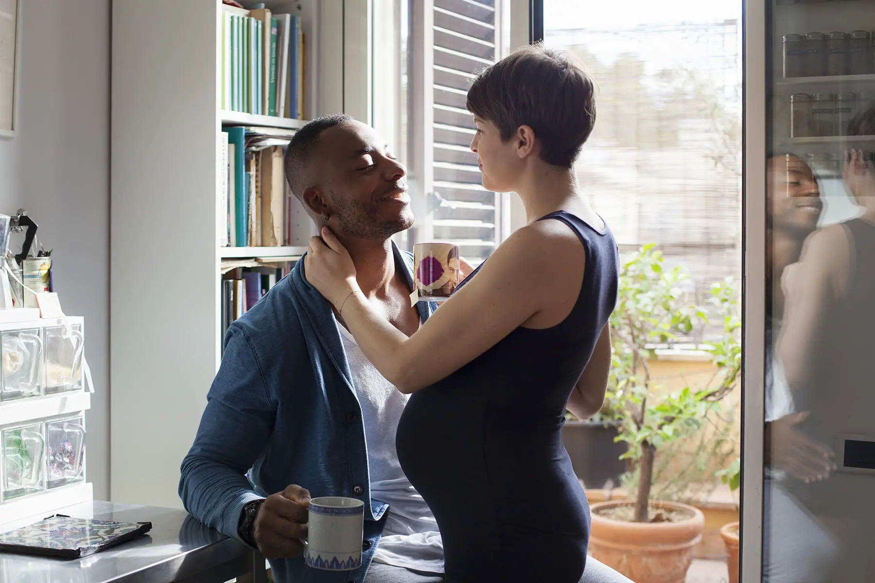 Smiling pregnant couple hugging while having tea together in the kitchen by an open balcony door.