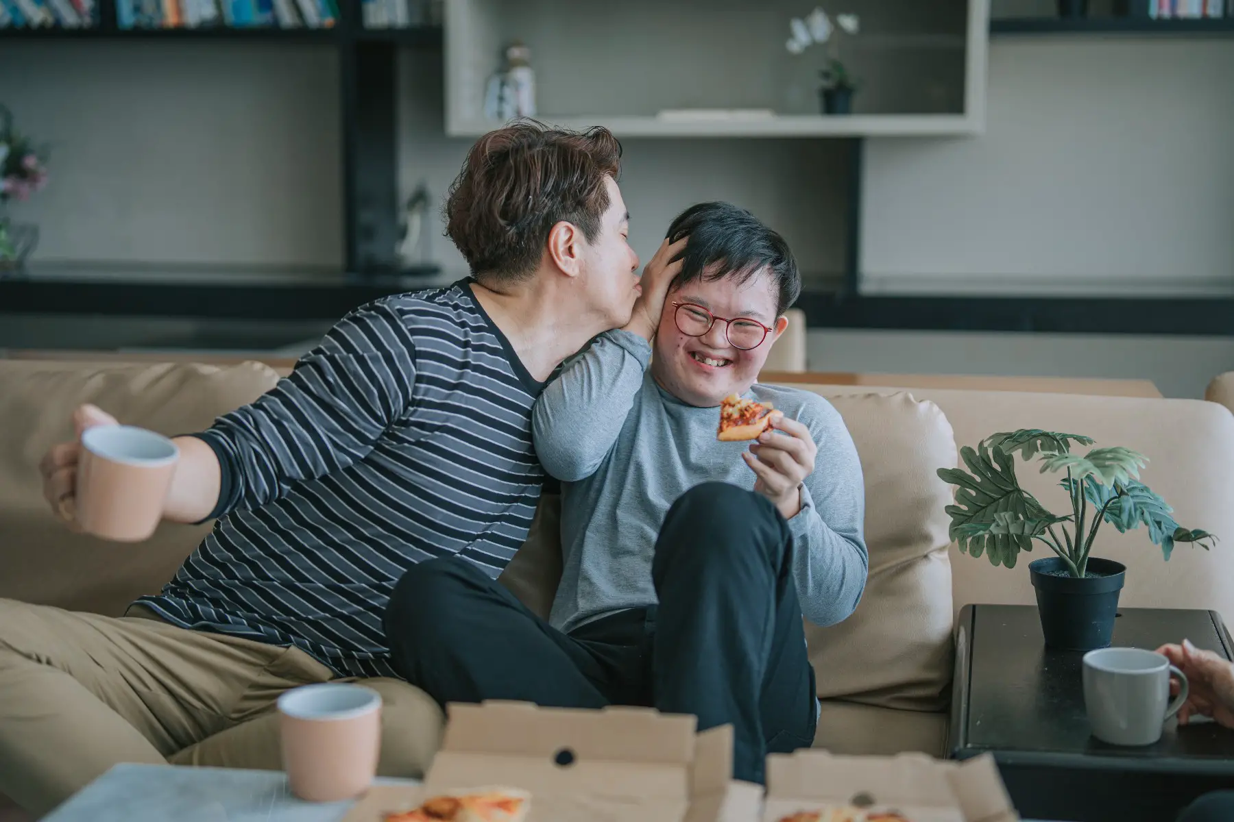Father leaning in to kiss his son with down syndrome, who is laughing and trying to avoid it. They are sitting on the sofa eating pizza.