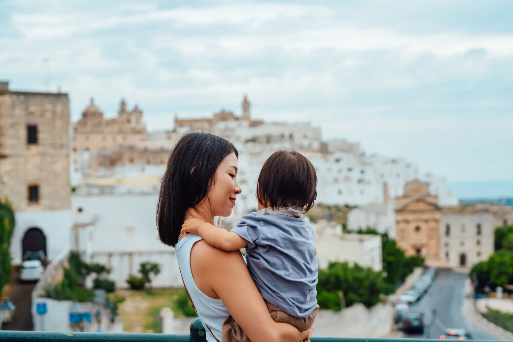 Young mother smiling lovingly at the toddler in her arms, with the old town of Ostuni (Italy) in the background.