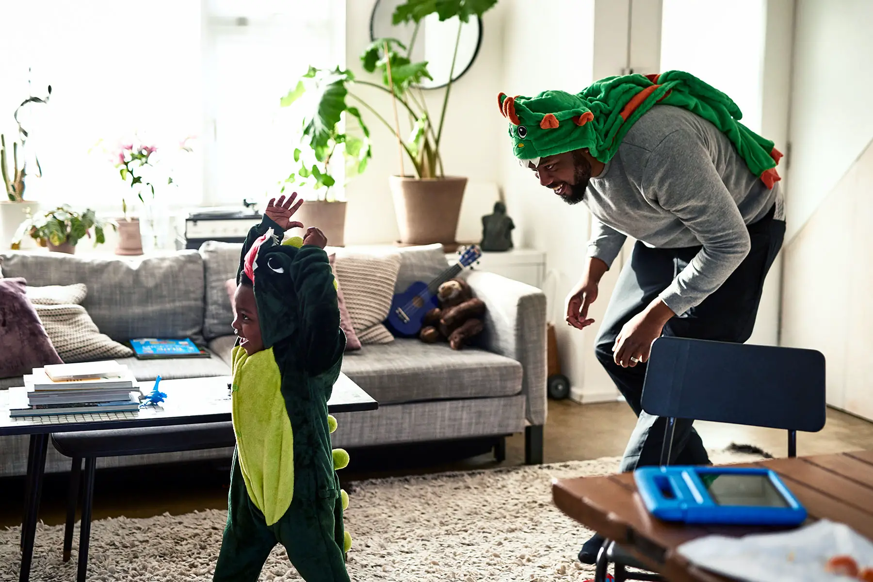 Young boy wearing dinosaur costume being chased by dad, half in costume, at home.