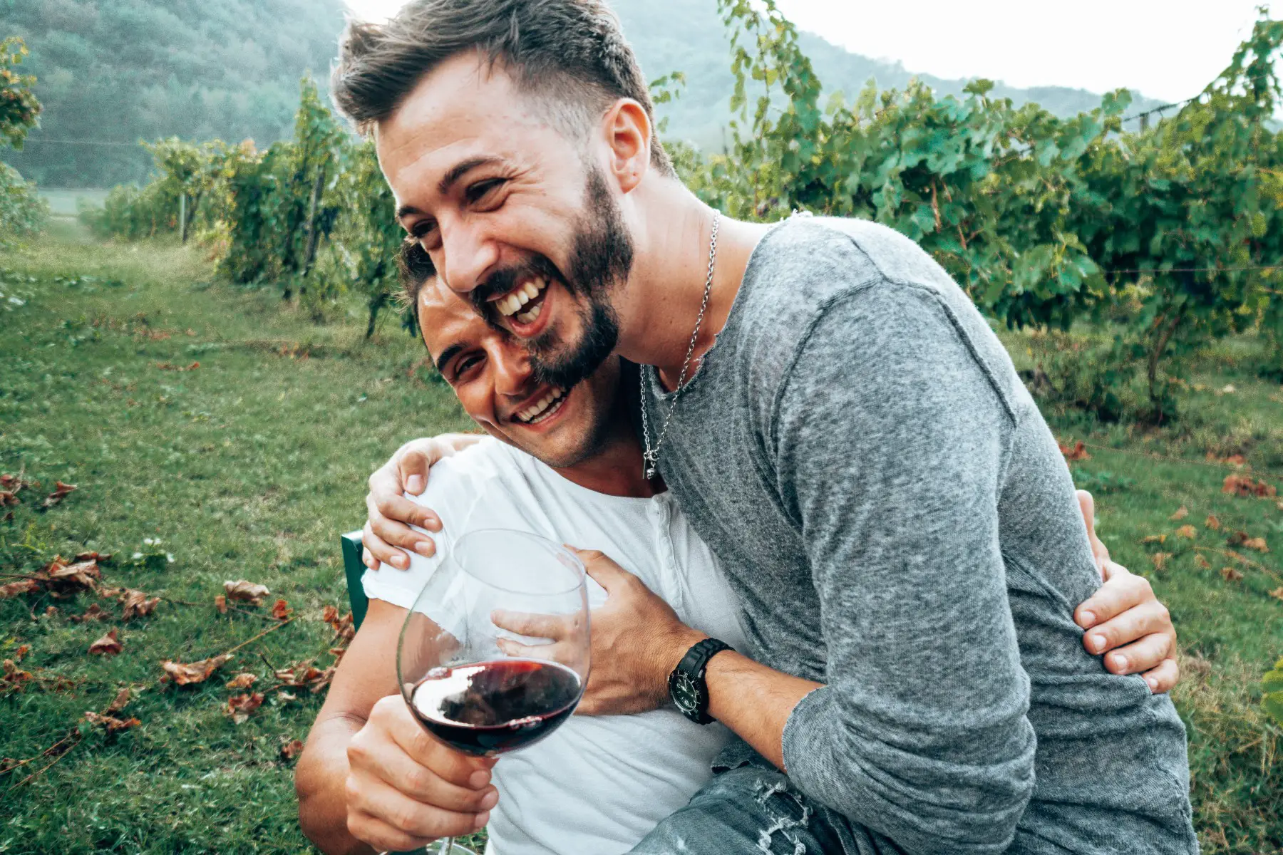 Gay couple laughing and toasting together during a wine tasting. One man is sitting on the other one's lap.